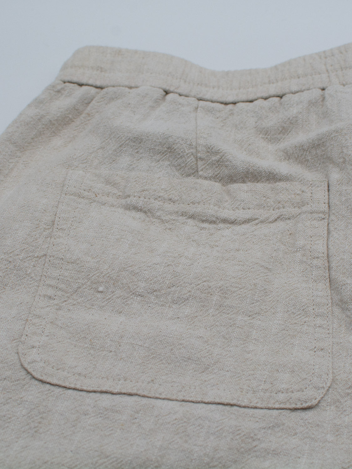 A close-up of the textured beige fabric from ID Menswears Phi Phi Linen Blend Drawstring Short. It highlights a single centered patch pocket with visible stitching, embodying the signature style of the 6 inseam shorts.