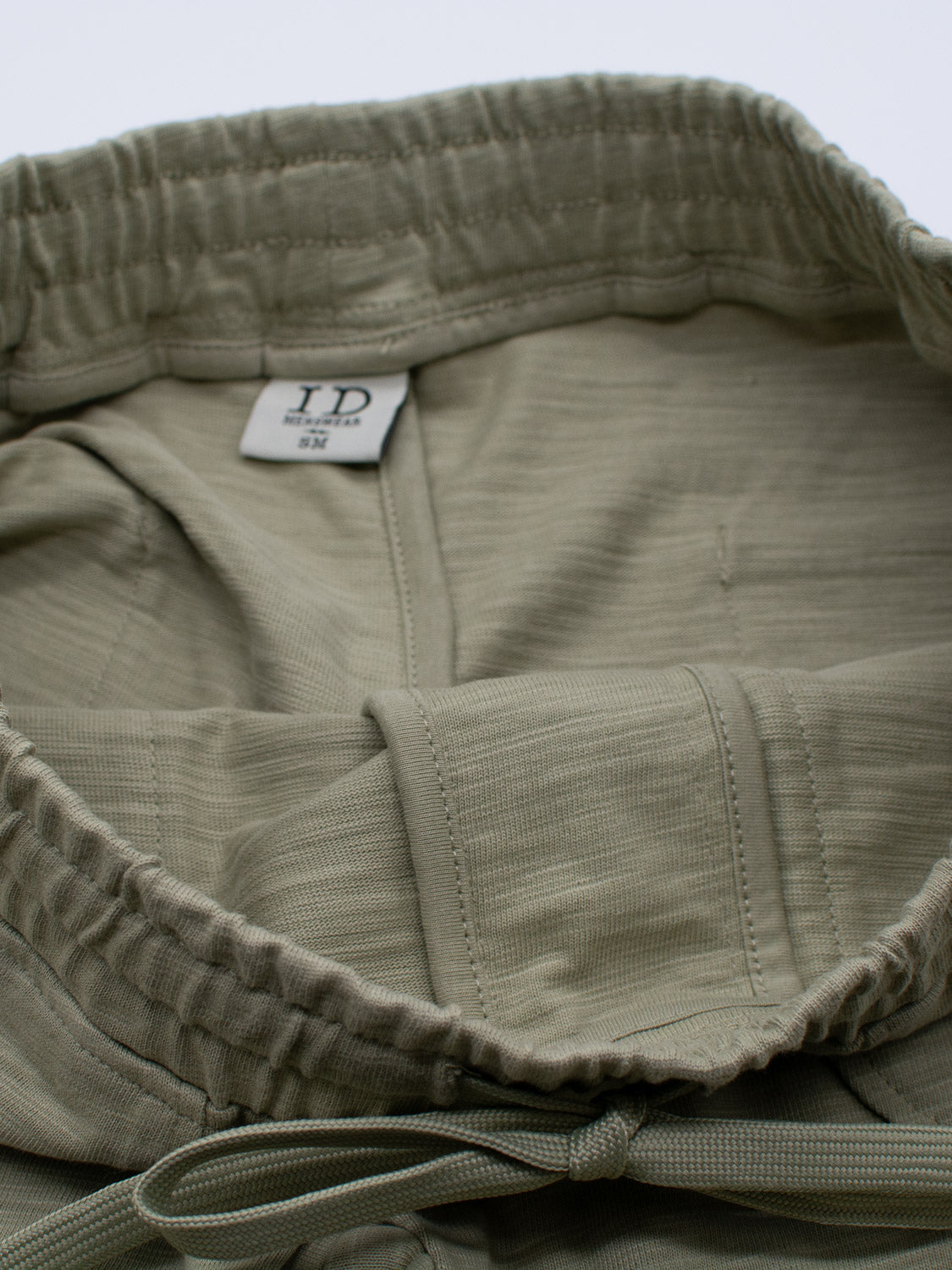 The Kahala Slub Cotton Drawstring Short by ID Menswear is shown in olive green with a relaxed fit, elastic waistband, and drawstring. The inside tag indicates size S/M. Made from slub cotton, the fabric has a subtle textured appearance.