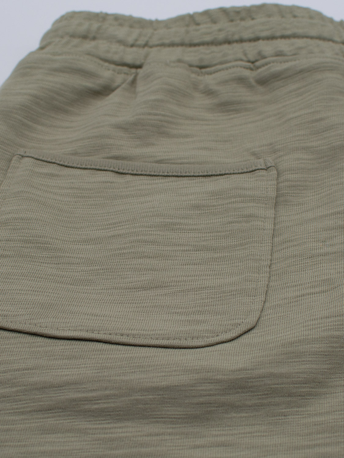 The Kahala Slub Cotton Drawstring Short by ID Menswear features a close-up of olive green fabric, crafted from slub cotton with a textured horizontal weave. These shorts have a relaxed fit, an elastic waistband thats partially visible at the top, and include a single patch pocket on the back.