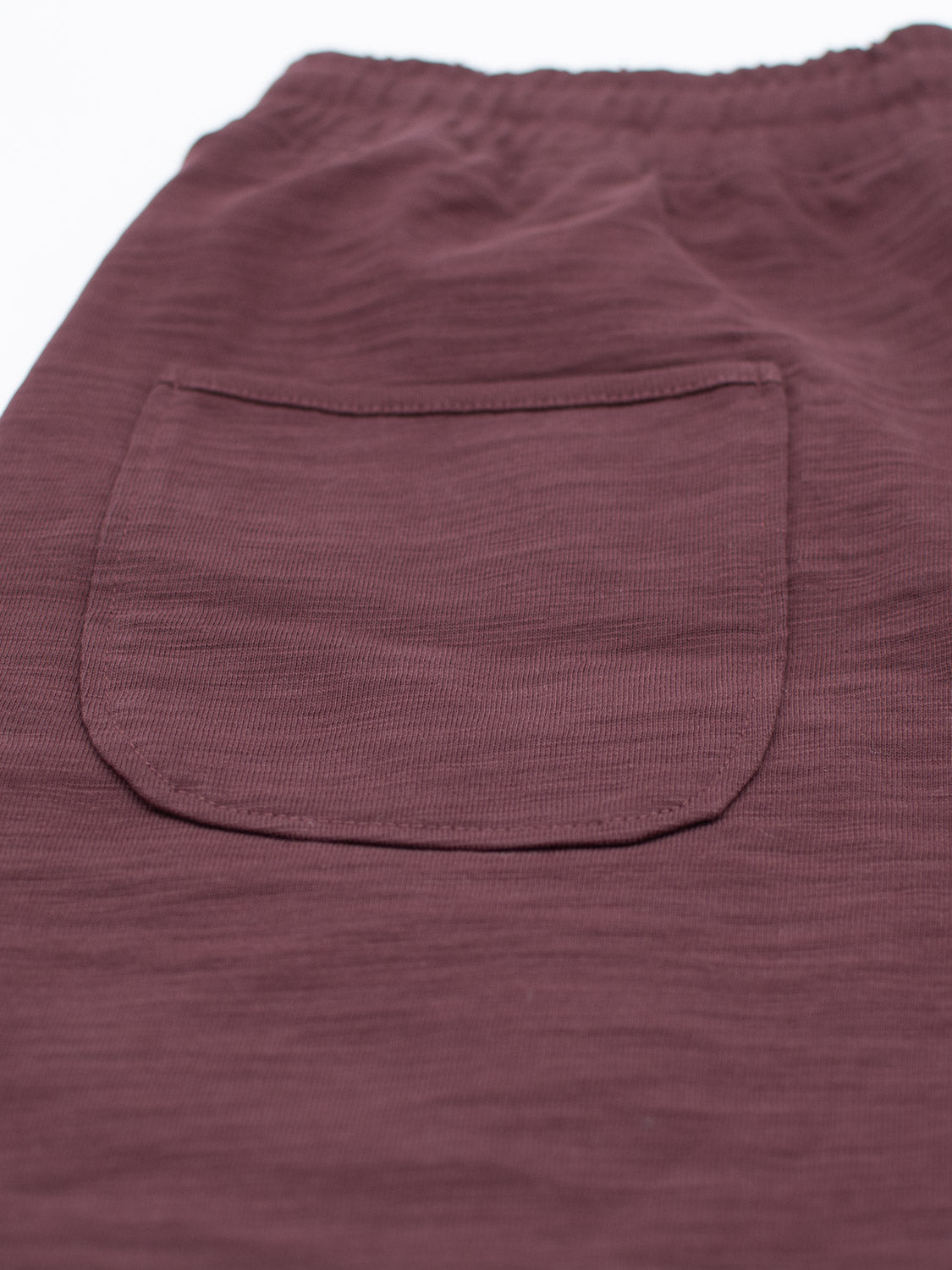 Close-up of the Kahala Slub Cotton Drawstring Short by ID Menswear, featuring maroon fabric with a back pocket. Its relaxed fit showcases a soft weave pattern. The slightly raised sewn-on pocket and slub cotton texture enhance the elastic waistband.