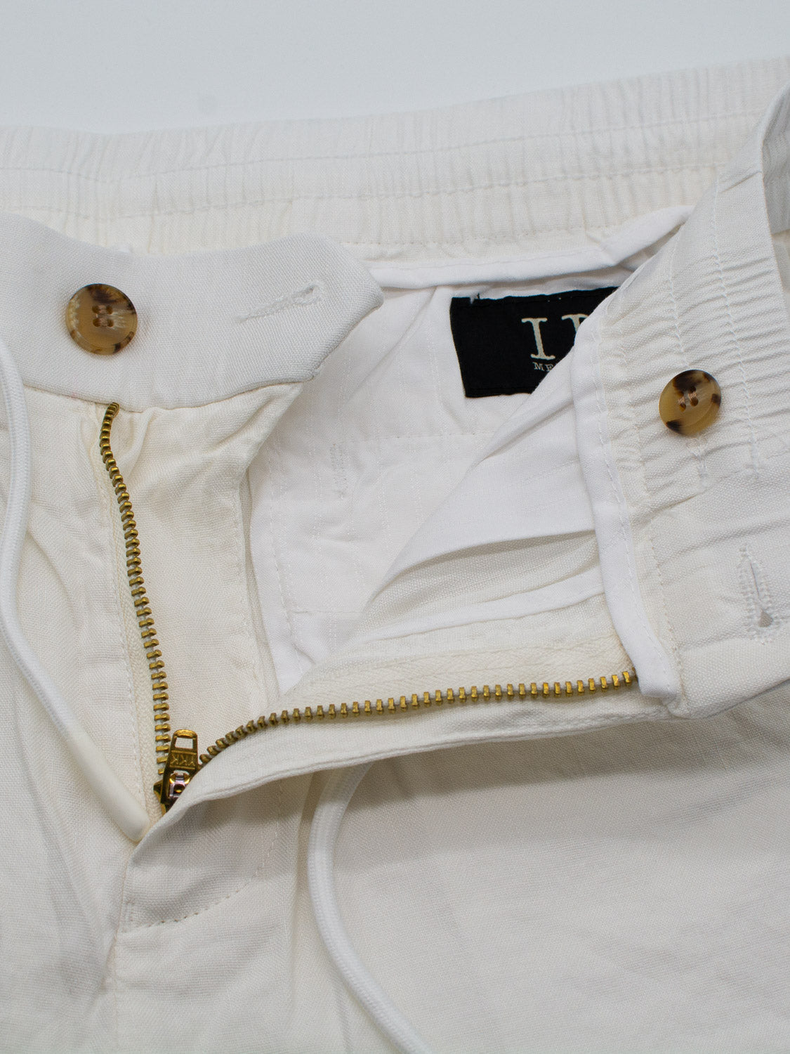 Close-up of the Driggs Linen Blend Short by ID Menswear in off-white, featuring a brass zipper, two light brown marbled waistband buttons, an internal black label, and a white drawstring—ideal for staying comfortable in warm weather.