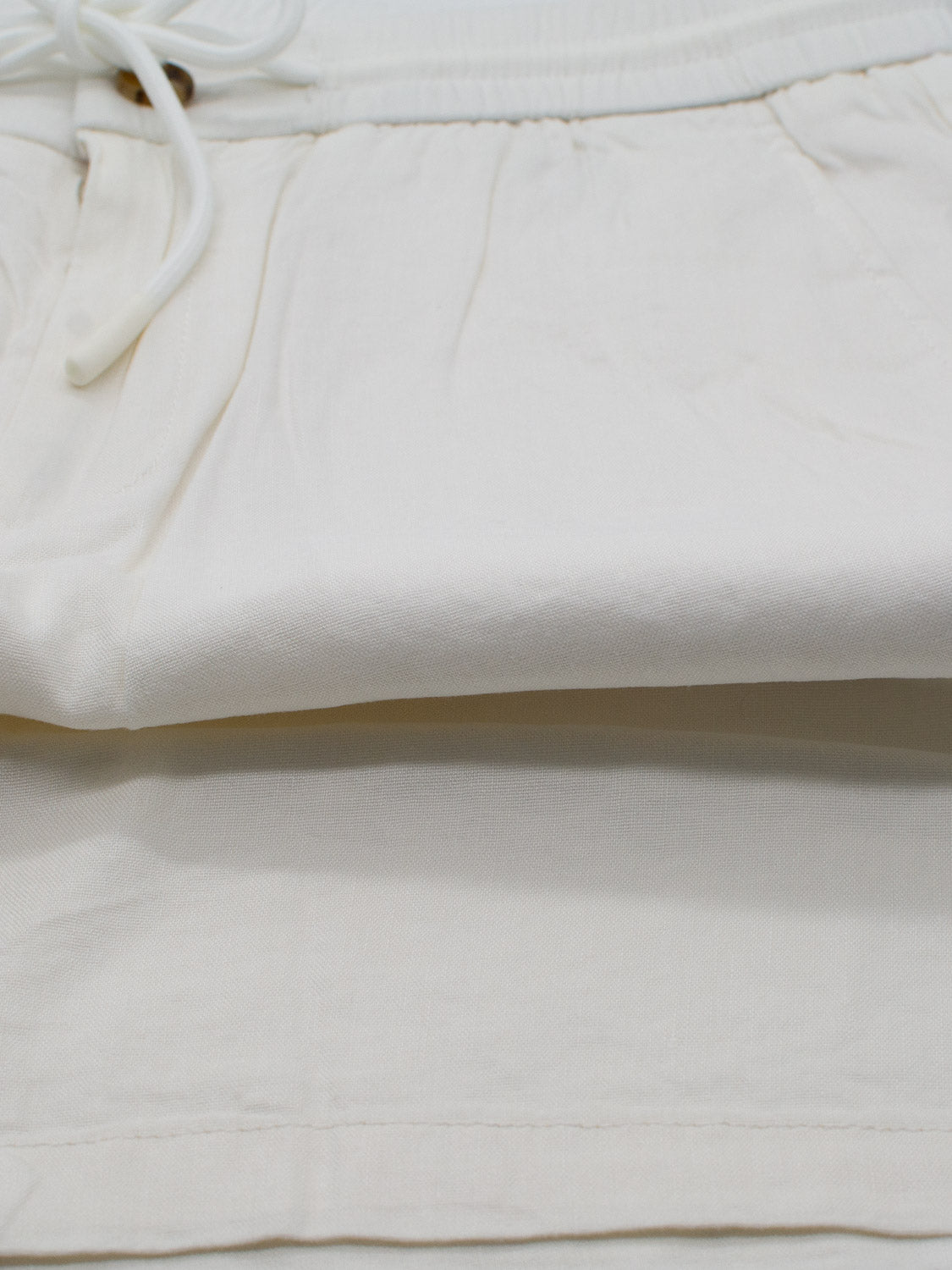 The Driggs Linen Blend Short by ID Menswear is folded neatly on a white surface. Ideal for warm weather, the off-white fabric looks soft and lightweight, featuring an elastic waistband and tied drawstring on the left side.