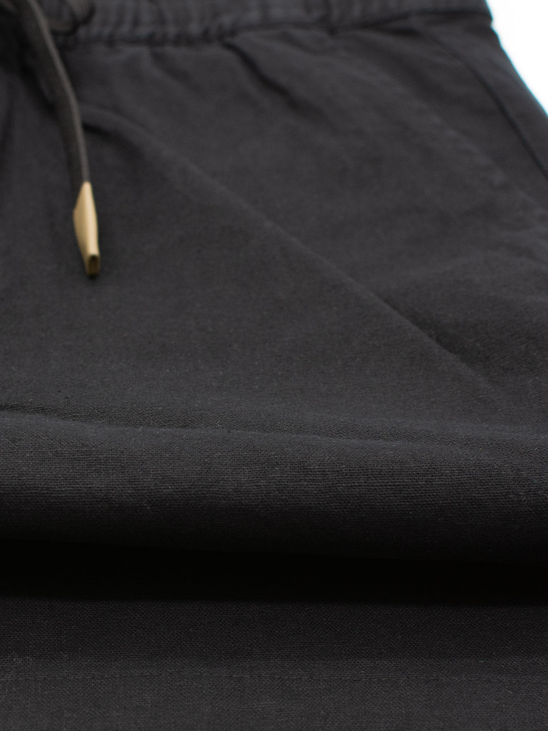 Close-up of the Kotao Linen Blend Drawstring Short by ID Menswear, featuring a black fabric with a drawstring and gold metal tip. The folded, textured material highlights the summer shorts elegance against a softly focused background.