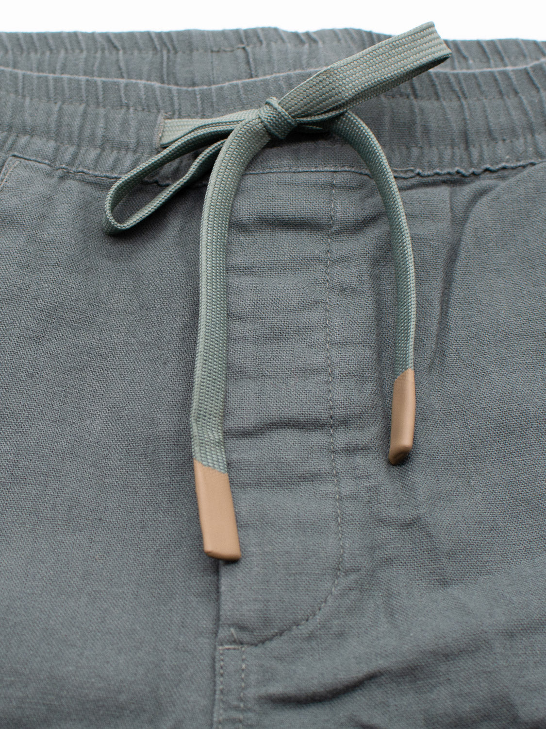 Close-up of ID Menswears Kotao Linen Blend Drawstring Shorts in grayish-green with an elastic waistband and drawstring featuring plastic tips. Visible seams with detailed stitching add to the look. Soft and comfortable, theyre ideal warm-weather bottoms.