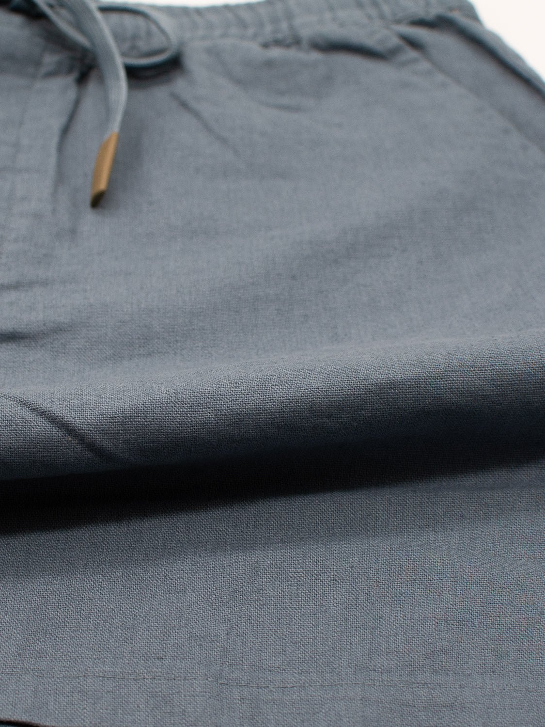 The image shows a close-up of the soft, textured gray-blue fabric of the Kotao Linen Blend Drawstring Short by ID Menswear. A seam and drawstring with metallic ends are visible, suggesting a comfortable summer fit.