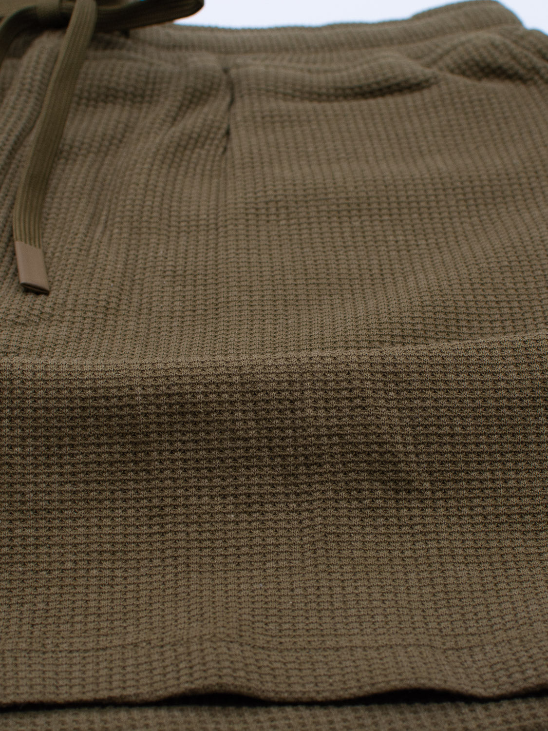 Close-up of Walker Waffle Knit Short by ID Menswear, showcasing a textured olive-green fabric with a subtle checkered pattern and a tied strap, possibly hinting at eco-friendly hemp material.