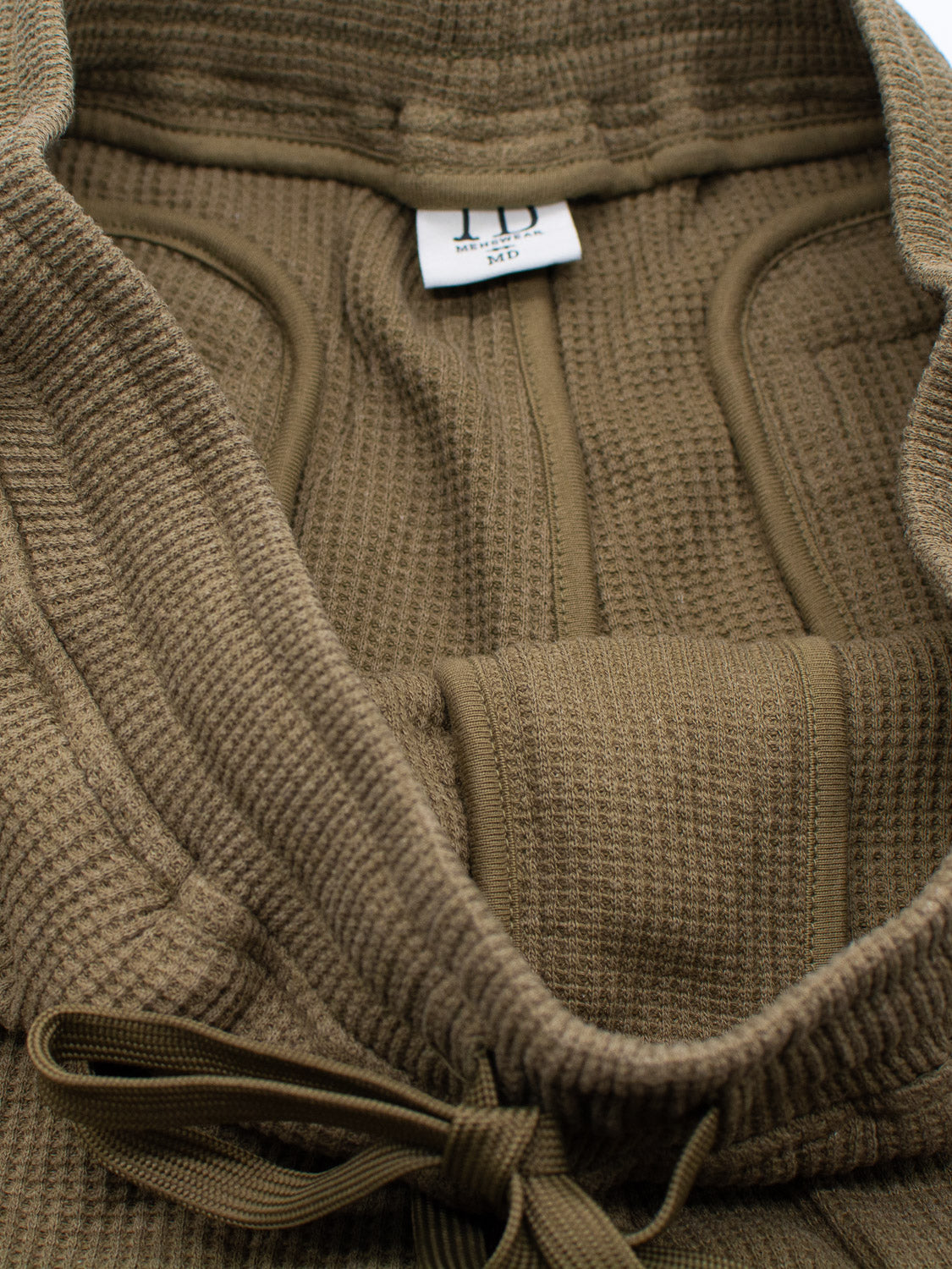 Close-up of a textured, olive-green hoodie with visible seams and drawstring, paired perfectly with Walker Waffle Knit Short by ID Menswear. A label inside reads 100 and 3470. The fabrics waffle-knit pattern gives both pieces a cozy yet rugged appearance.