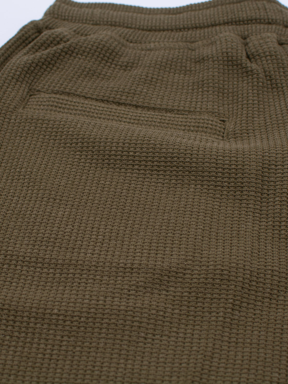 A close-up of the Walker Waffle Knit Short by ID Menswear features soft, cozy organic cotton with an olive green waffle pattern. The focus is on a slightly gathered-edge pocket, making these shorts ideal for casual wear or lounging.