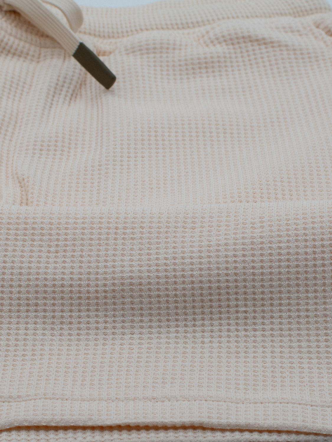 A close-up of the Walker Waffle Knit Short by ID Menswear shows a beige waffle-textured fabric, likely organic cotton, featuring a drawstring threaded through a metal eyelet.