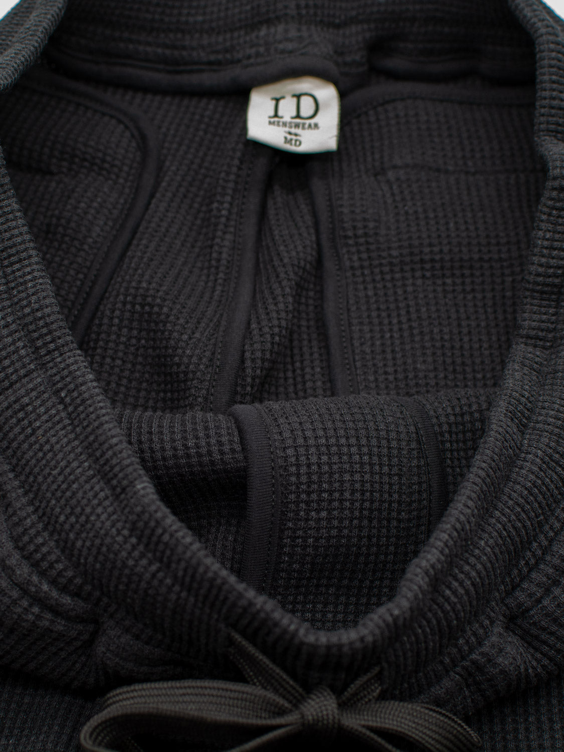 A black waffle-knit hooded garment from ID Menswear features a visible label and is paired with matching Walker Waffle Knit Shorts for a cohesive look. The hood has a black tie, showcasing the intricate fabric texture prominently.