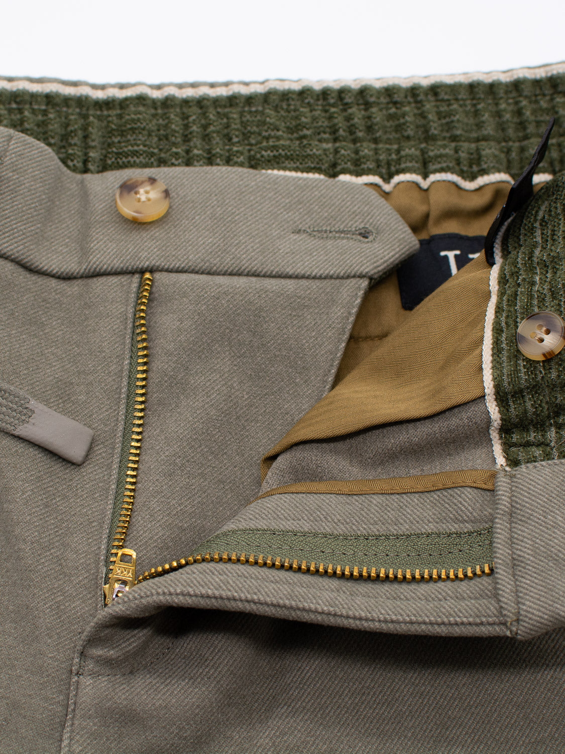 Close-up of ID Menswears Brighton Brushed Polyester Rayon Pant in khaki, featuring a gold zipper slightly open to show olive green fabric and a speckled green waistband. These drawstring pants include brown buttons and a tag with partially visible letters.
