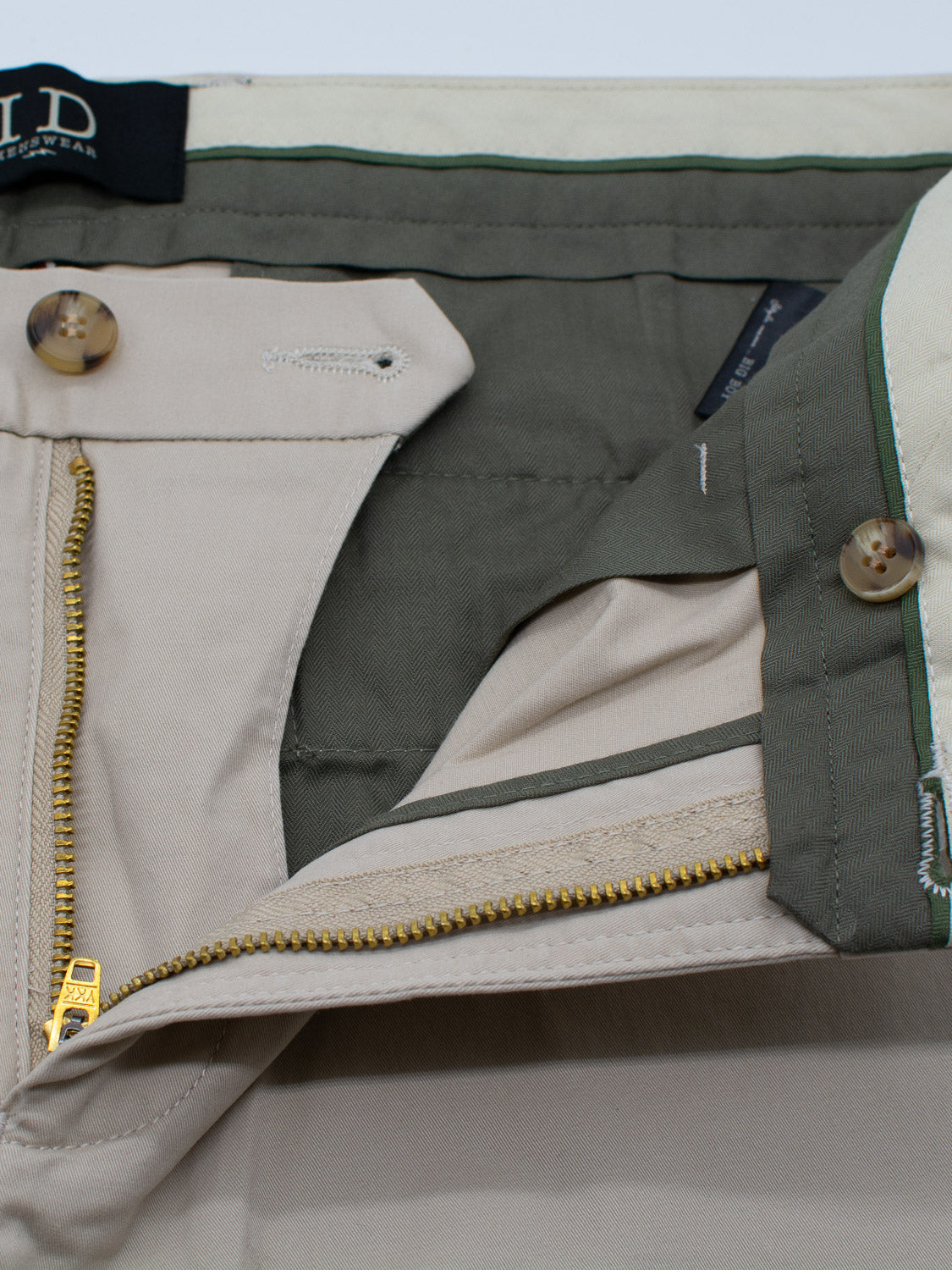 Close-up of beige Bigboy Relaxed Flooded Chino by ID Menswear, featuring a green-lined interior, a partially open brass zipper and button closure. The relaxed, baggy fit adds to its lightweight flexibility with interior waistband buttons and attached clothing tag.