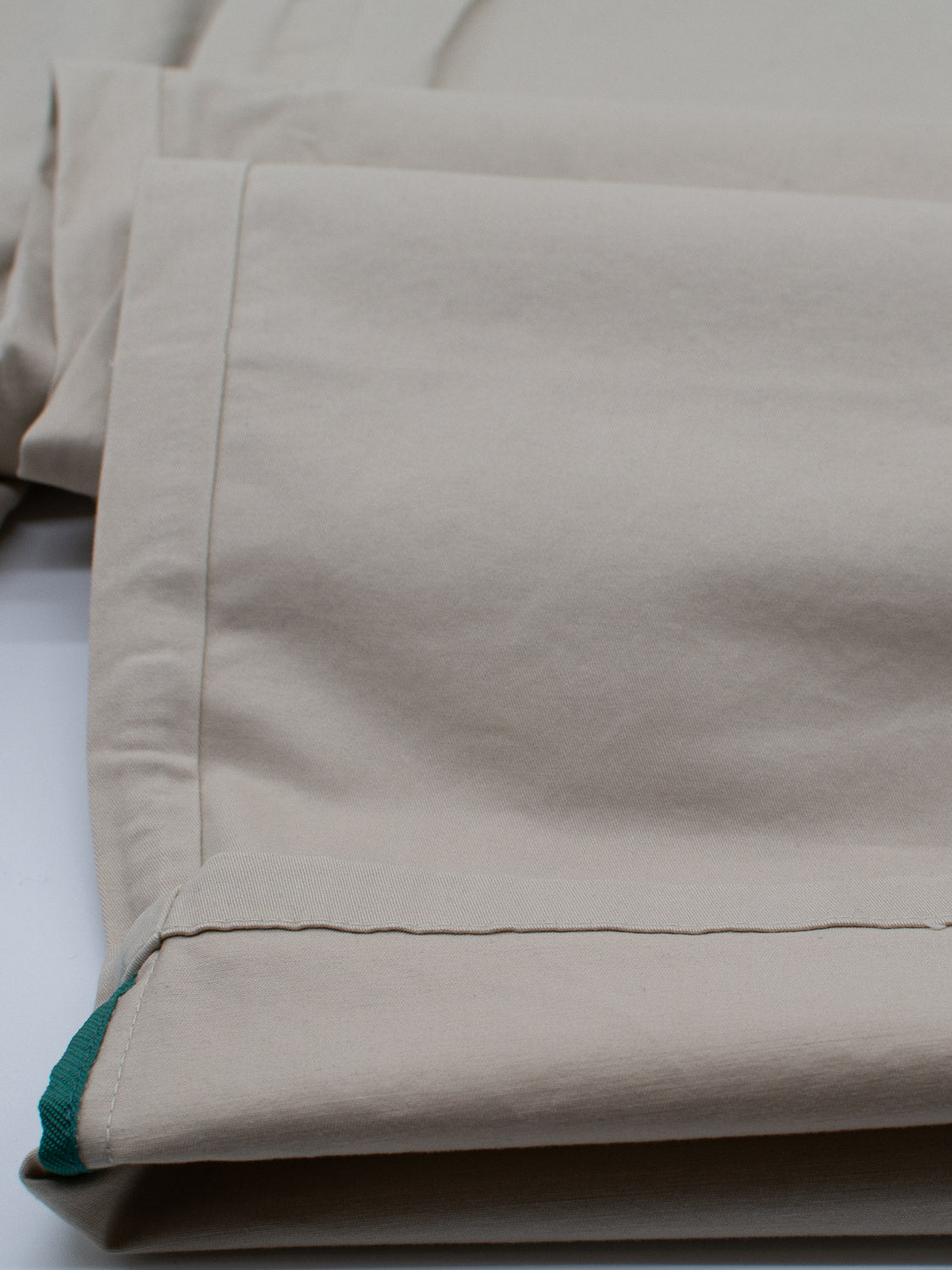 A close-up of the Bigboy Relaxed Flooded Chino by ID Menswear showcases neatly folded beige fabric with visible stitching and lightweight flexibility. A hint of green fabric peeks out, evoking a relaxed and baggy fit, resting effortlessly on a smooth white surface.