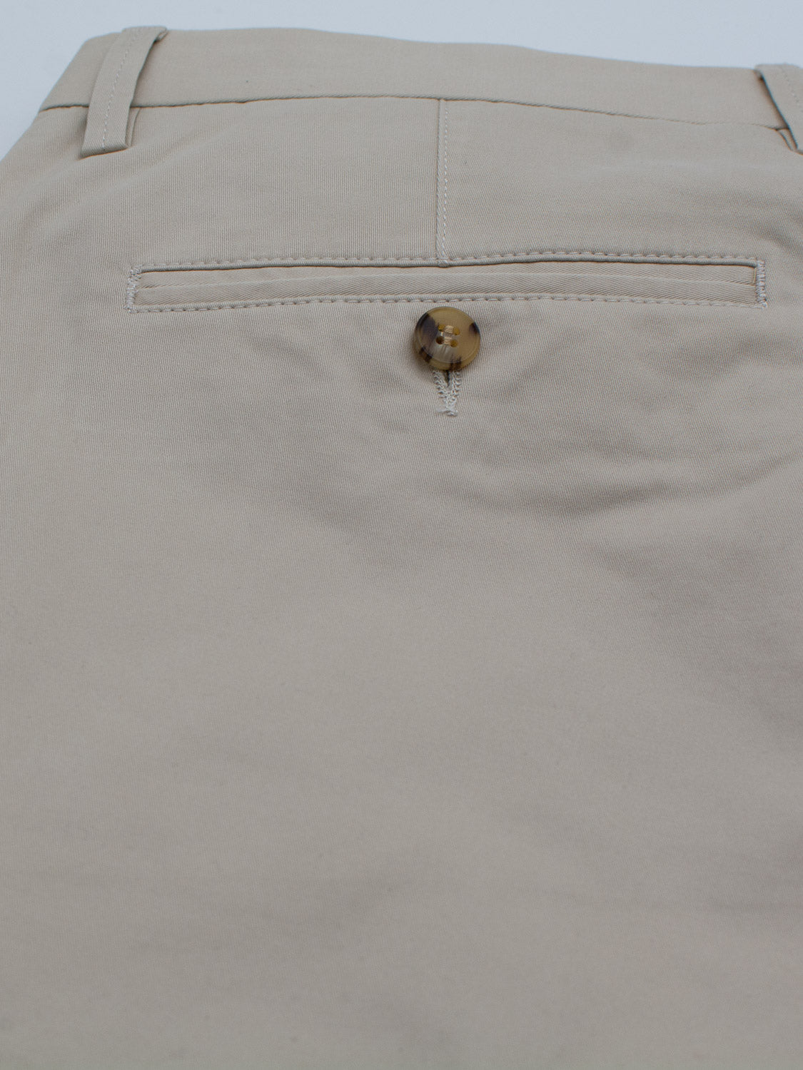 A close-up showcases the back pocket of beige Bigboy Relaxed Flooded Chino pants by ID Menswear, featuring a brown and cream button closure. The lightweight fabric appears smooth and well-pressed, emphasizing the neatly sewn pocket line.