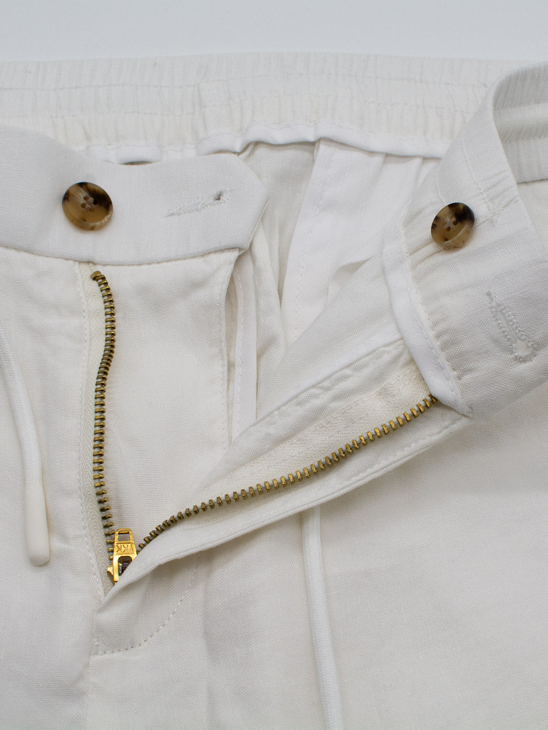 Close-up of cream-colored, lightweight Bedford23 Linen Blend Drawstring Pant by ID Menswear, featuring a partially open gold zipper and tan buttons. It has an elastic waistband with visible drawstring ends beside the stylish zipper.