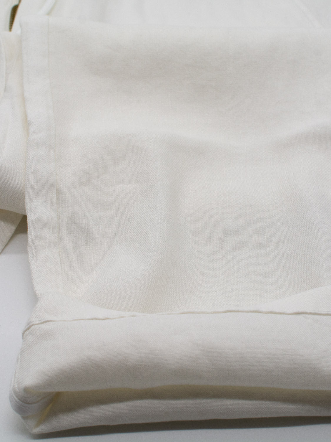A neatly folded piece of off-white fabric with a smooth texture lies on a flat surface, reminiscent of ID Menswears lightweight and crisp Bedford23 Linen Blend Drawstring Pant, with subtle shadows highlighting its folds against a soft white background.