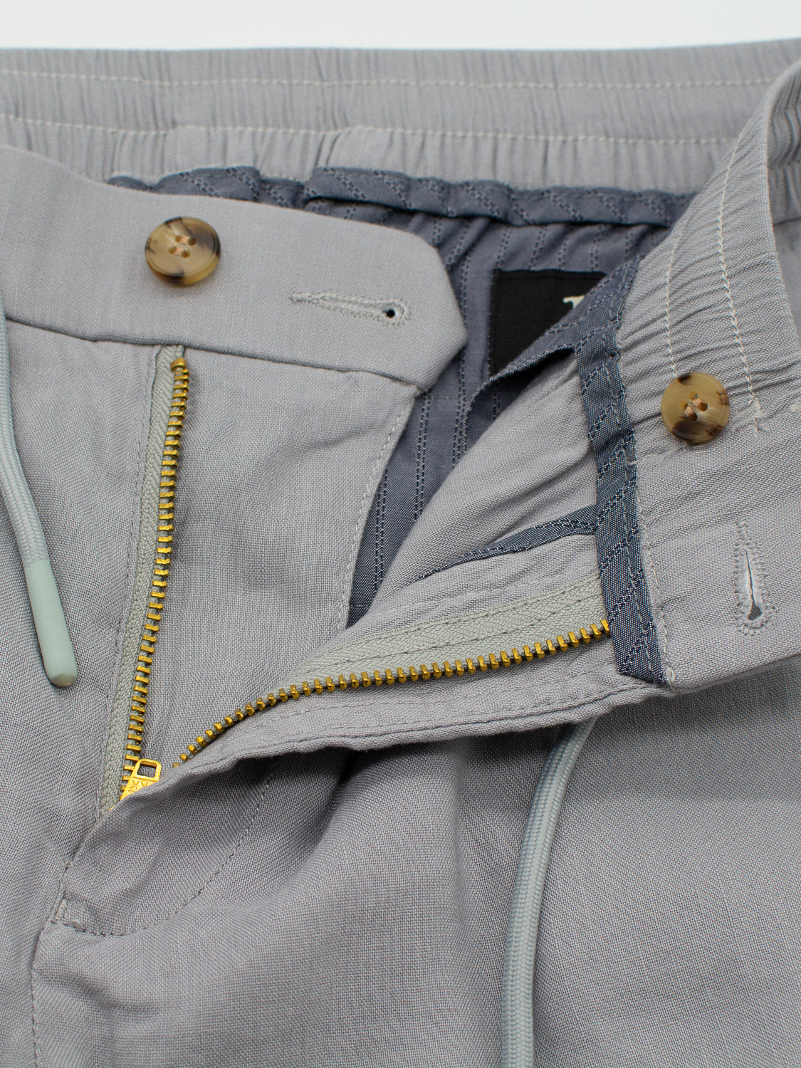 Close-up of the Bedford23 Linen Blend Drawstring Pant by ID Menswear, highlighting its gray elastic drawstring waistband with green-tipped cords. Features include a gold zipper, two brown buttons, and a darker gray interior lining for lightweight comfort.