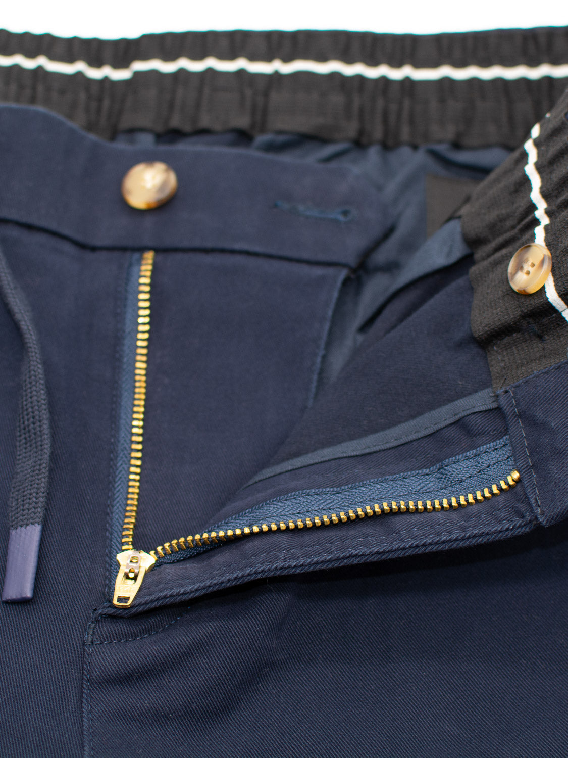 The Dyker Cotton Twill Drawstring Jogger by ID Menswear features a navy blue design with an open gold zipper, visible brown buttons, and black-and-white striped waistband. Crafted for durability, the cotton twill fabric showcases standout seams and texture while offering style and comfort.