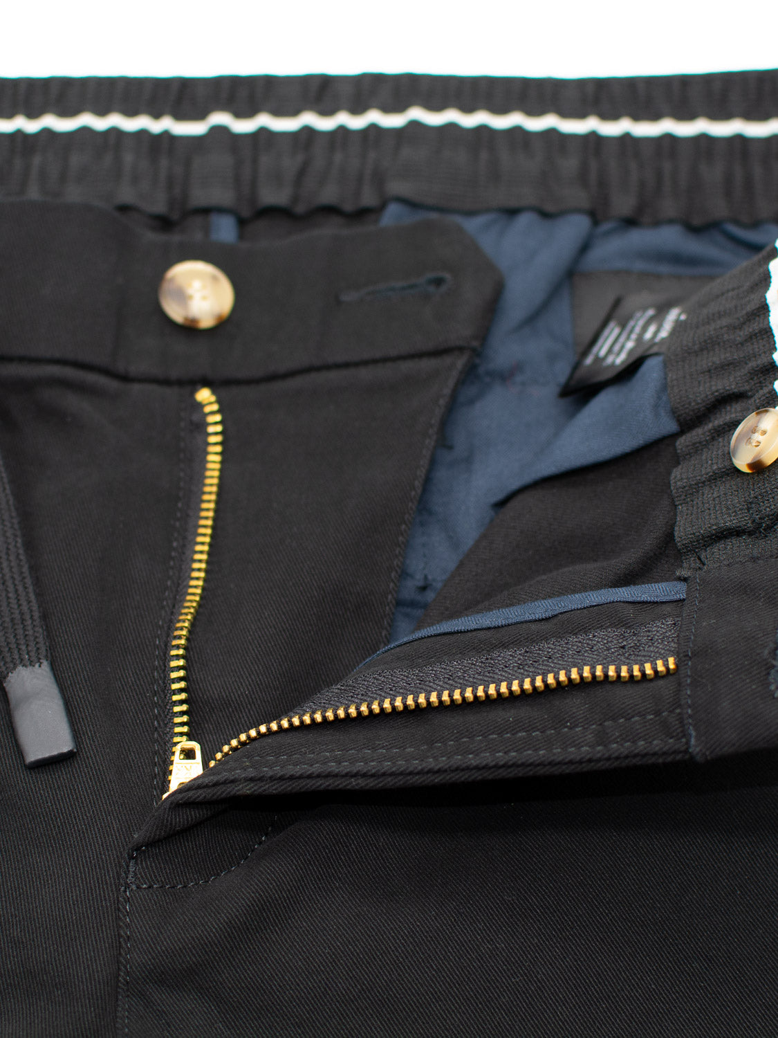 Close-up of the Dyker Cotton Twill Drawstring Jogger by ID Menswear, showcasing an open black zipper and button with gold accents. The inner blue fabric lining is revealed, along with an elastic waistband suggestive of joggers and a partially visible tag.