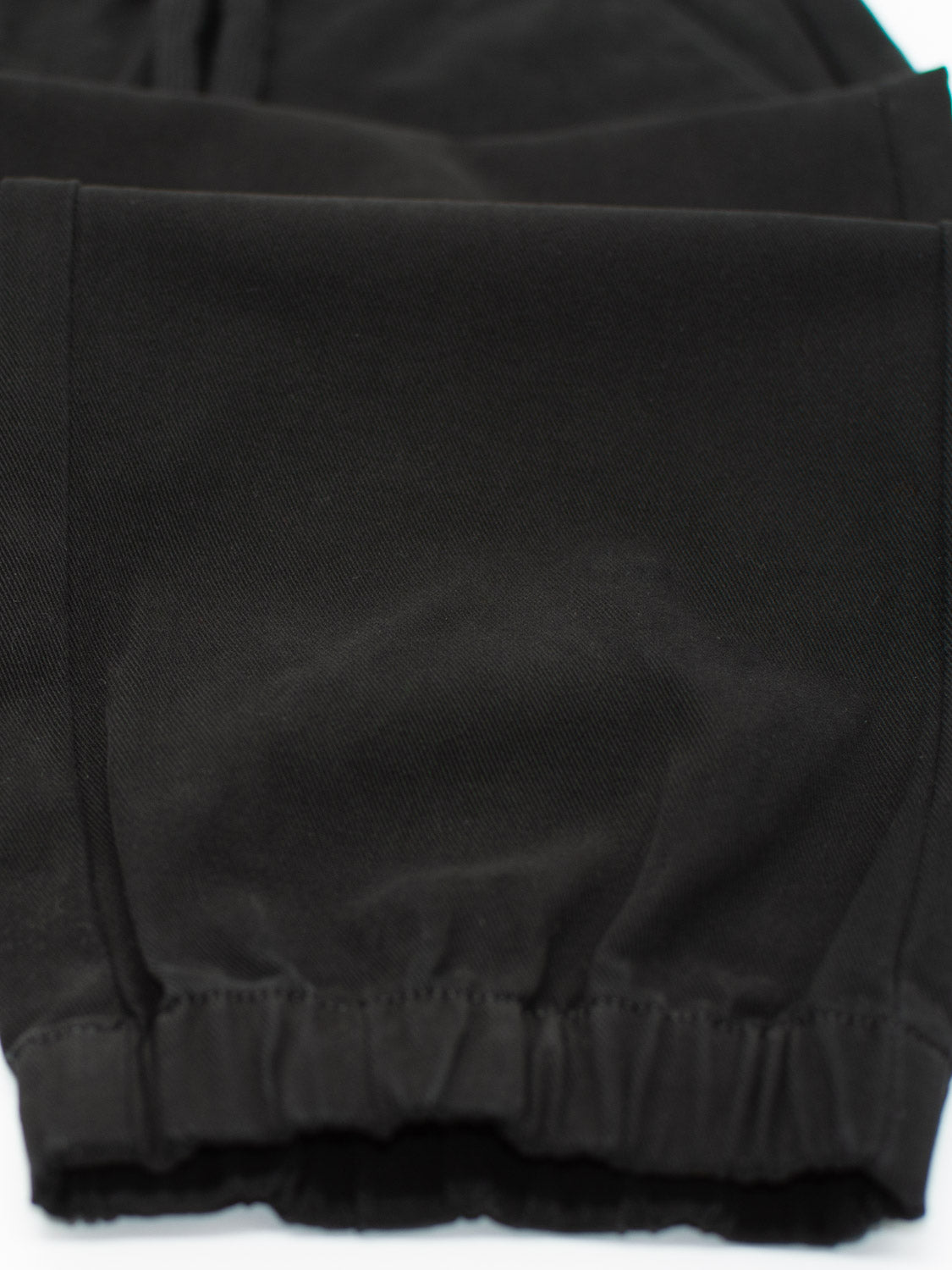 Zoom in on the ID Menswear Dyker Cotton Twill Drawstring Jogger, featuring a black hue with elastic waist, hem, and cuffs. The fabric boasts a smooth, slightly shiny texture and visible stitching highlights the exquisite detail.