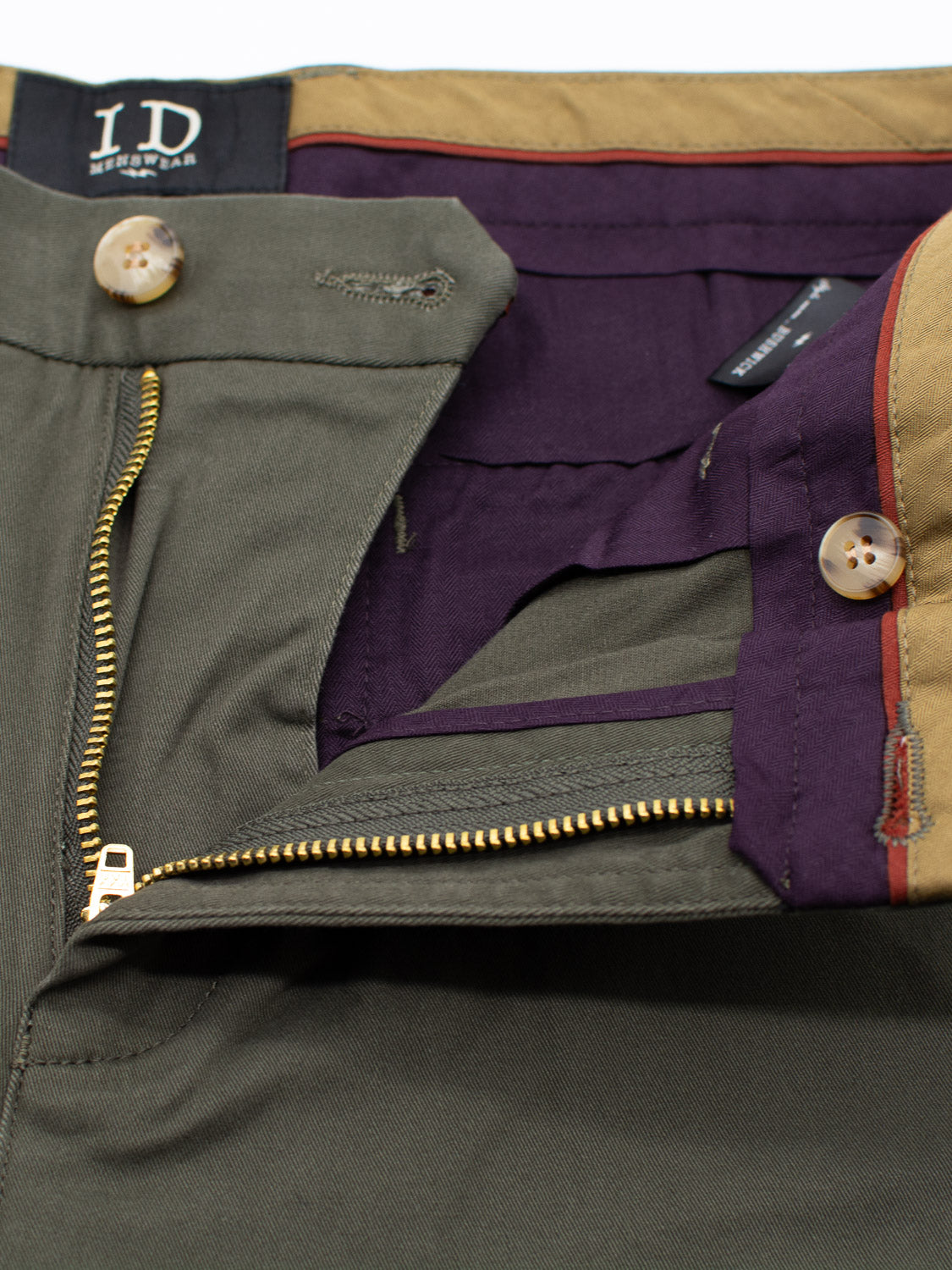 Close-up of the Bushwick Twill Stretch Chino pants by ID Menswear, highlighting the waistband and open zipper. These relaxed-fit trousers are made from olive green cotton twill with a dark purple interior lining, brown trim, and buttons. The brand label is visible on the waistband.