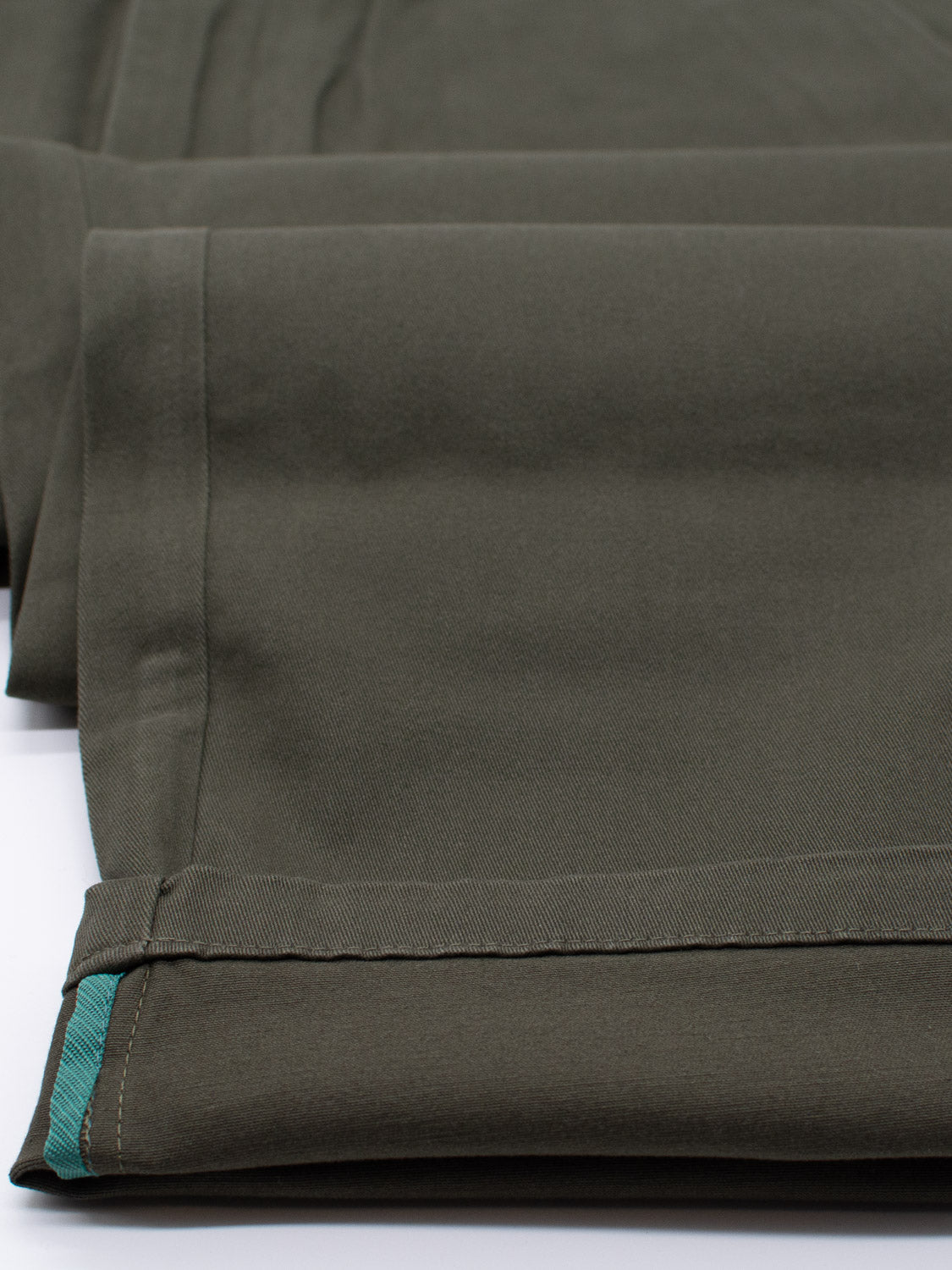 Close-up of neatly folded olive green cotton twill fabric with turquoise edge stitching, layered to highlight its texture and smoothness on a white background, reminiscent of ID Menswears Bushwick Twill Stretch Chino.