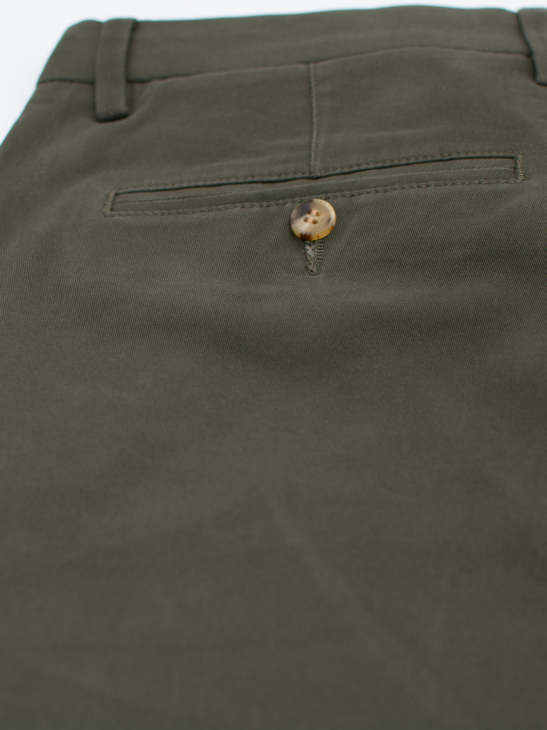 Close-up of ID Menswears Bushwick Twill Stretch Chino in olive green, highlighting the back pocket. These relaxed-fit pants feature a brown button with a light swirl pattern, and the smooth, neatly stitched cotton twill fabric showcases their quality.
