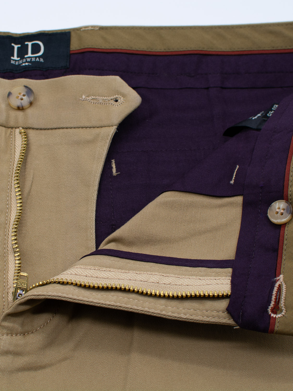 Close-up of beige Bushwick Twill Stretch Chino trousers from ID Menswear featuring a zipper and button closure. The inside waistband, lined with dark purple fabric, includes buttons and a tag with the brand initials. Made from soft cotton twill, these chinos offer style and comfort.