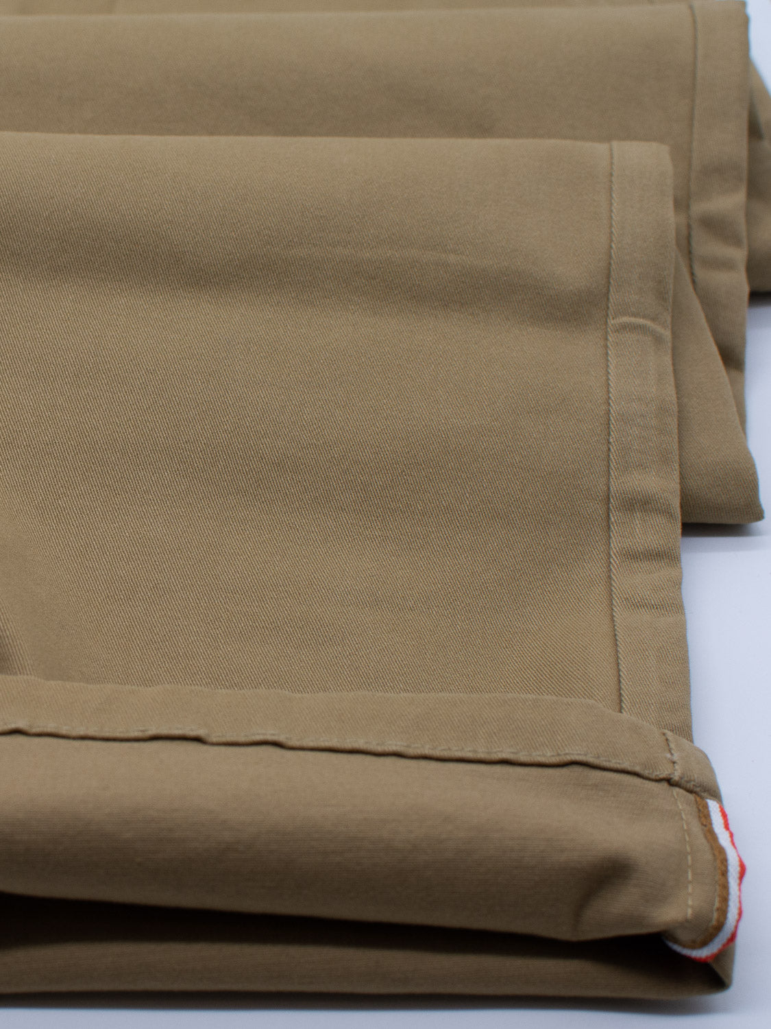 Close-up of tan folded fabric with visible stitching, arranged like a pair of ID Menswears Bushwick Twill Stretch Chino pants. Some edges reveal red and white accents. The smooth cotton twill fabric shows a slight texture.