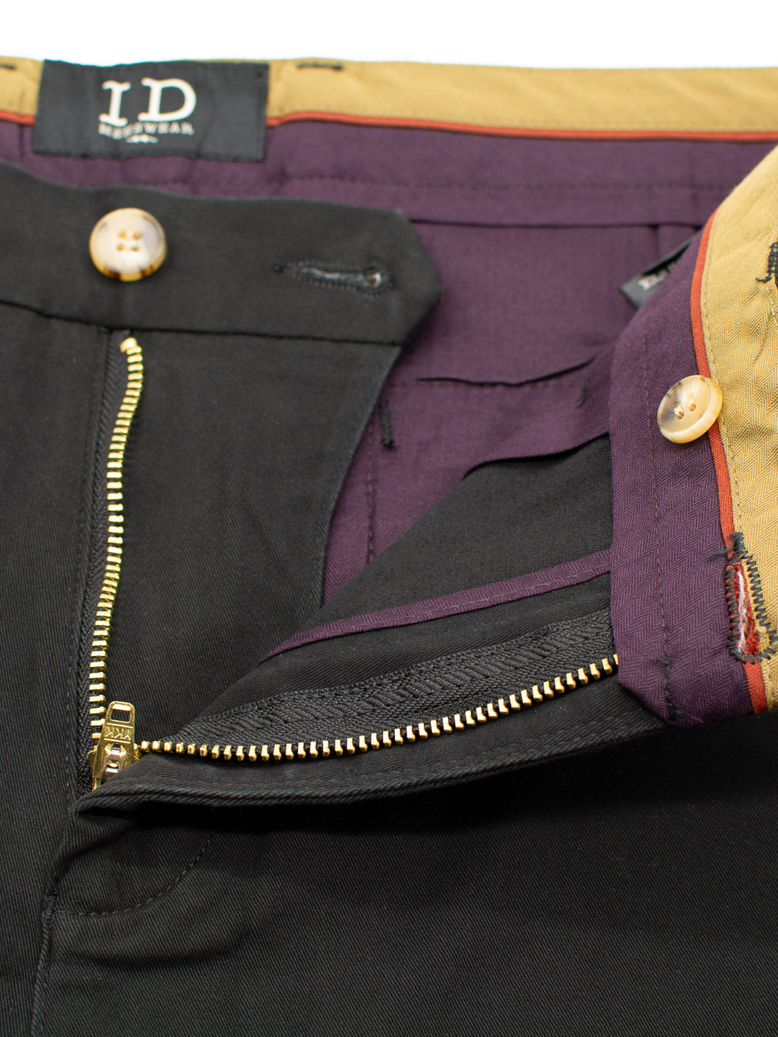 A close-up shows ID Menswears Bushwick Twill Stretch Chino, black relaxed-fit trousers with a partially open zipper. The waistband boasts contrasting tan and purple, adding flair, while golden buttons decorate the waist and fly. An I.D label is visible inside.