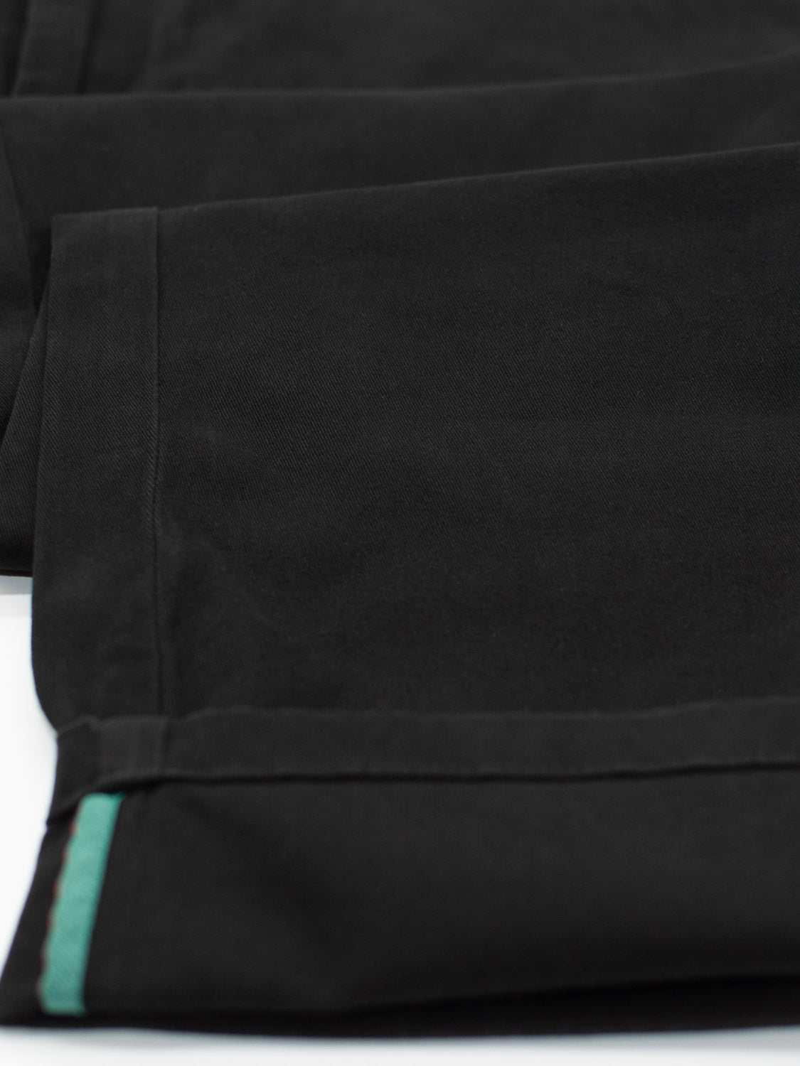 Close-up of the hem of relaxed-fit, black Bushwick Twill Stretch Chino trousers by ID Menswear, featuring a green trim. Made from cotton twill, the fabric looks smooth and neatly stitched, showcasing classic style.