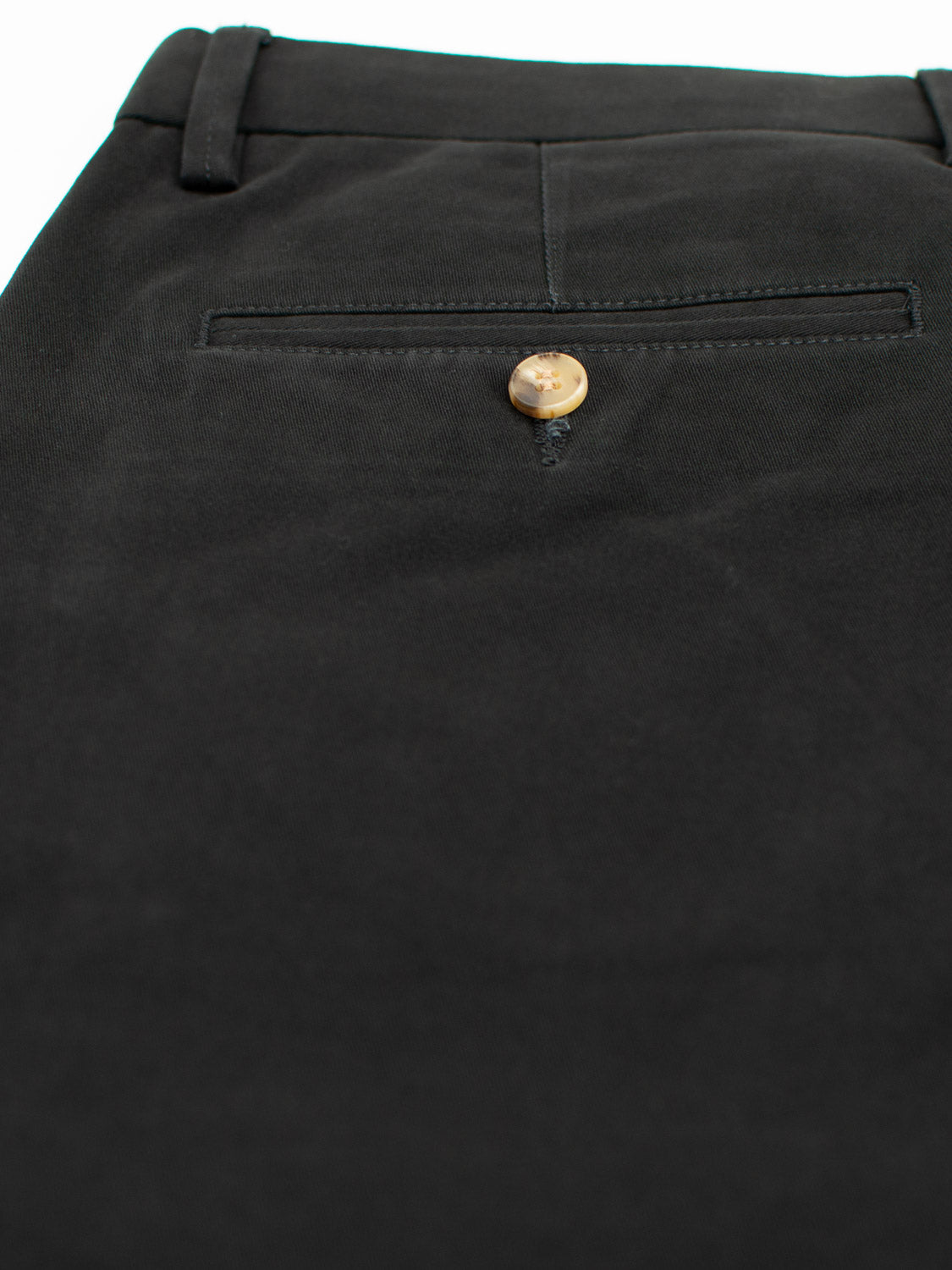 Close-up of black Bushwick Twill Stretch Chino pants by ID Menswear, showcasing a single back pocket with a light brown button. The smooth cotton twill fabric is slightly reflective, and the button subtly contrasts against the dark material.