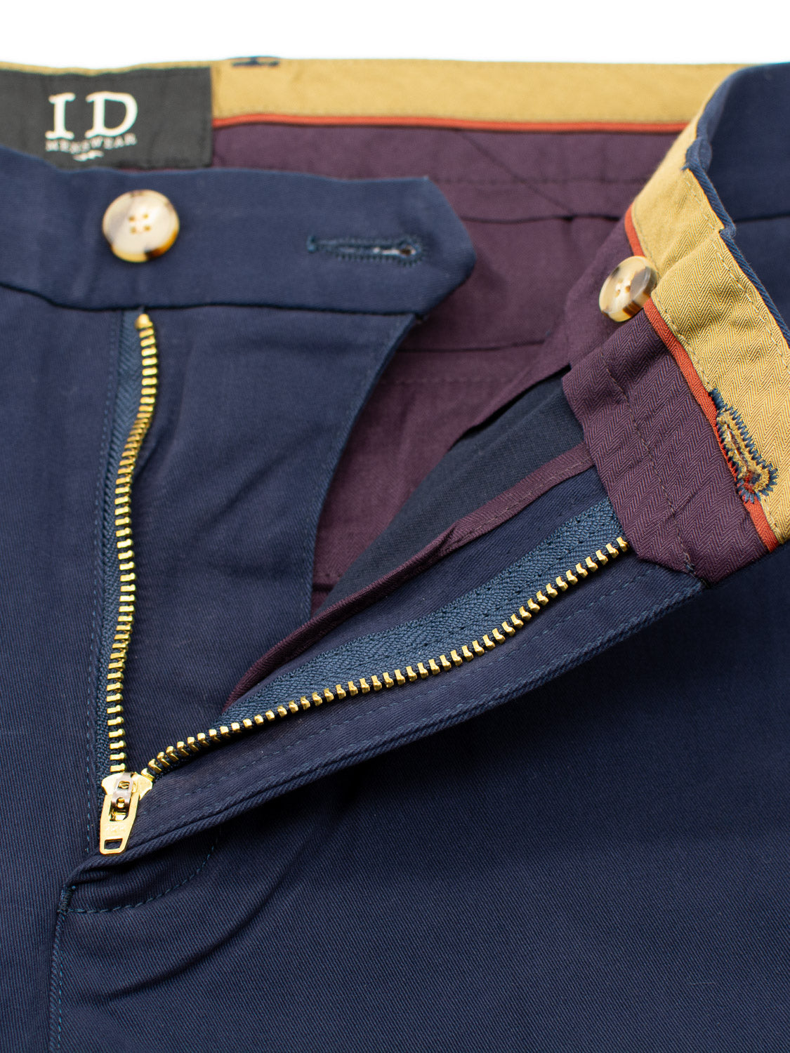 Close-up of navy blue relaxed-fit Bushwick Twill Stretch Chino by ID Menswear. It features a partially open zipper and tan buttons, with cotton twill inner lining showing burgundy fabric and a tan waistband. The visible ID label highlights its craftsmanship.