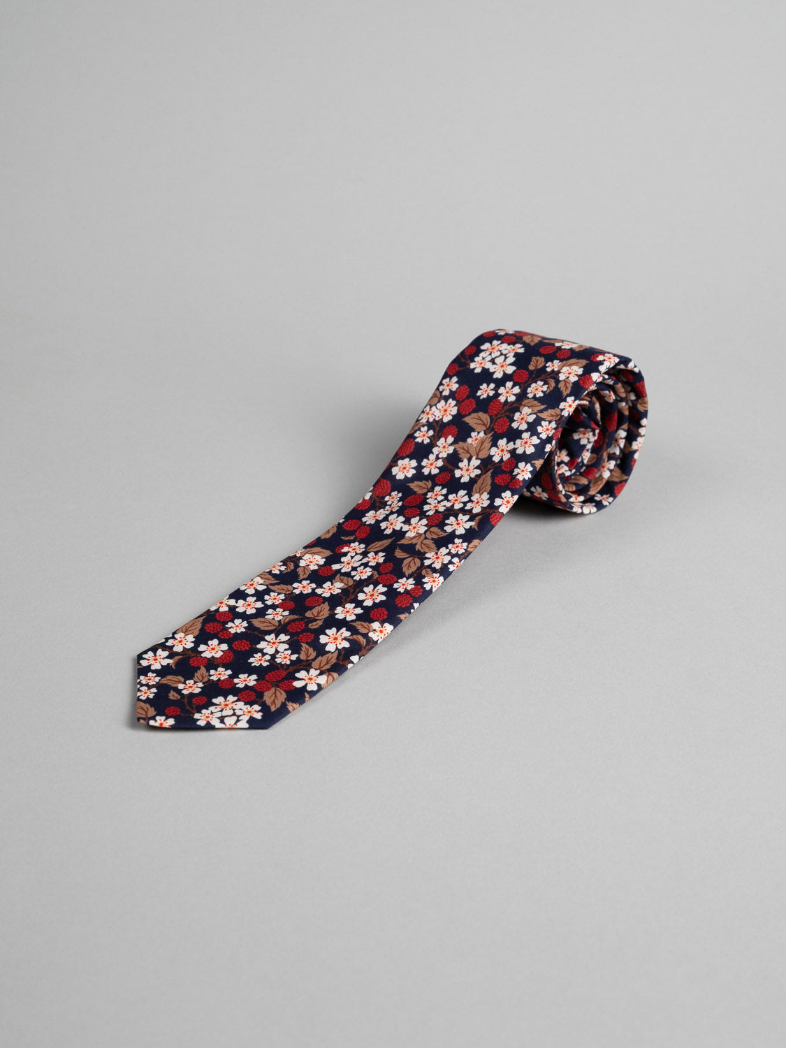 The ID Menswear Liberty of London Tana Lawn™ Karen Tie is handmade in Brooklyn, featuring a navy blue floral pattern with red, white, and brown flowers. Shown rolled on a light gray background.