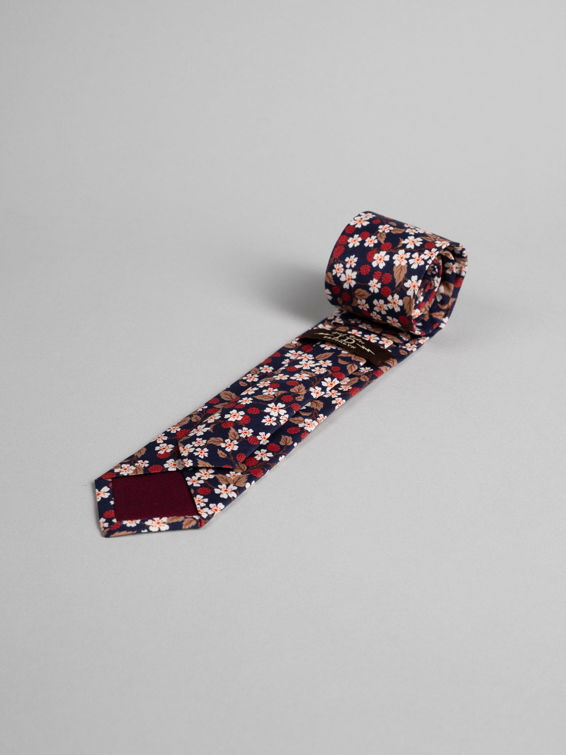 A Liberty of London Tana Lawn™ Karen Tie by ID Menswear, made from fine cotton with a dark red floral pattern, is shown rolled on a light gray surface—a distinctive handmade Brooklyn accessory.