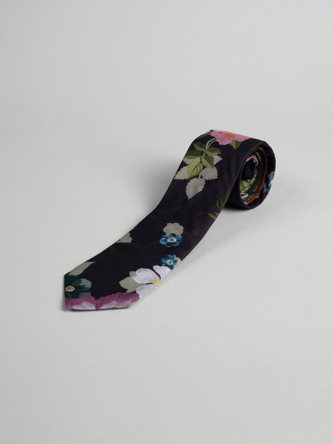 The Liberty of London Tana Lawn™ Nysa Tie by ID Menswear is handmade in Brooklyn, featuring a black base with green leaves and purple, blue, and white flowers on soft cotton, shown rolled on a light gray background.