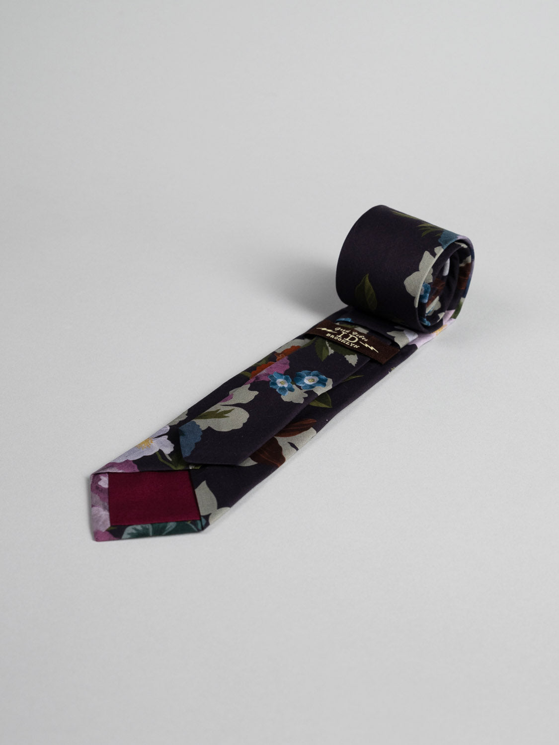 The Liberty of London Tana Lawn™ Nysa Tie by ID Menswear, handmade in Brooklyn from soft cotton with a dark floral design, is shown neatly rolled on a plain light gray background.