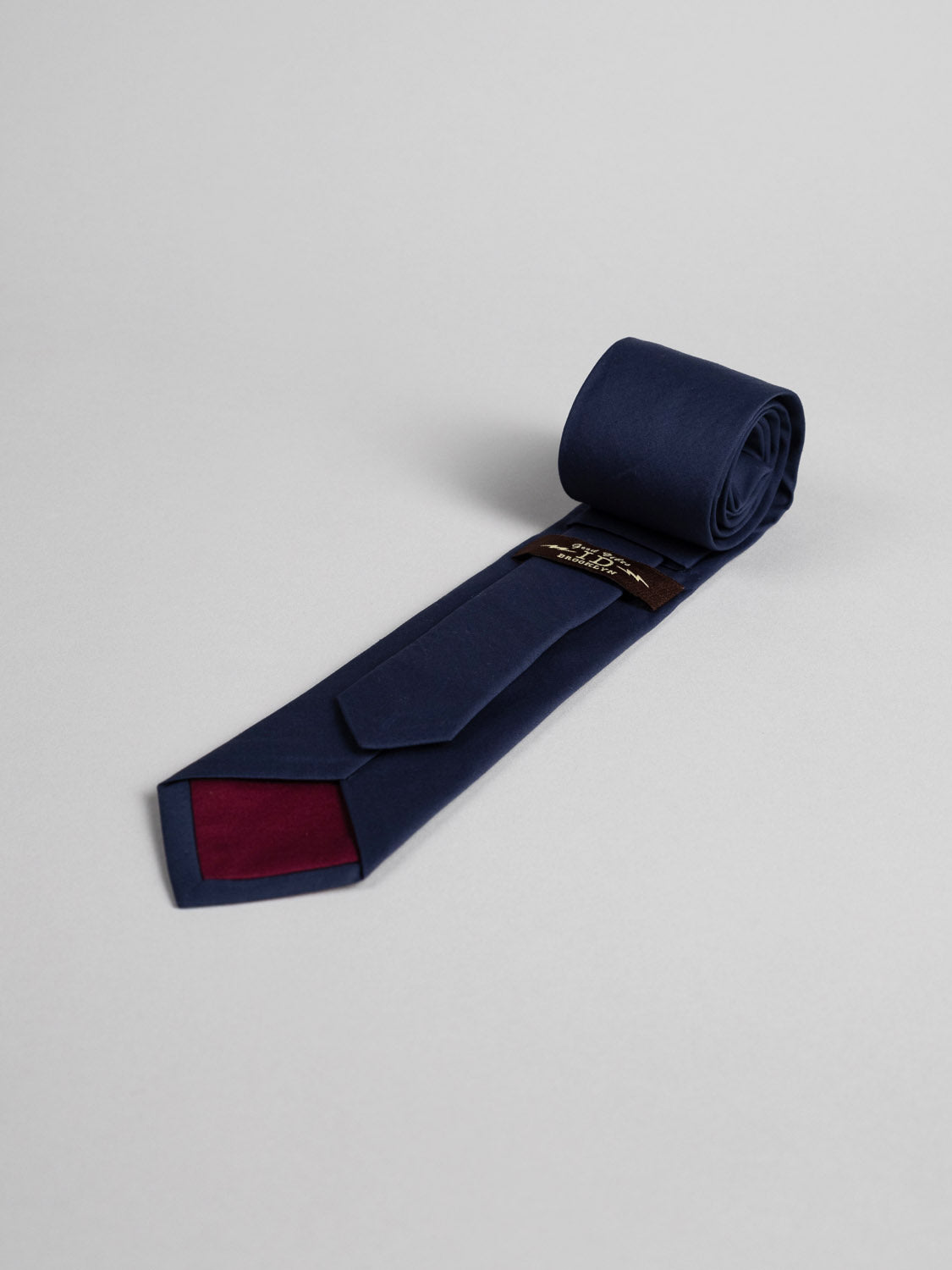The Liberty of London Tana Lawn™ Navy Tie by ID Menswear, handmade in Brooklyn with a maroon triangle detail on the reverse, is shown rolled up on a light gray background.