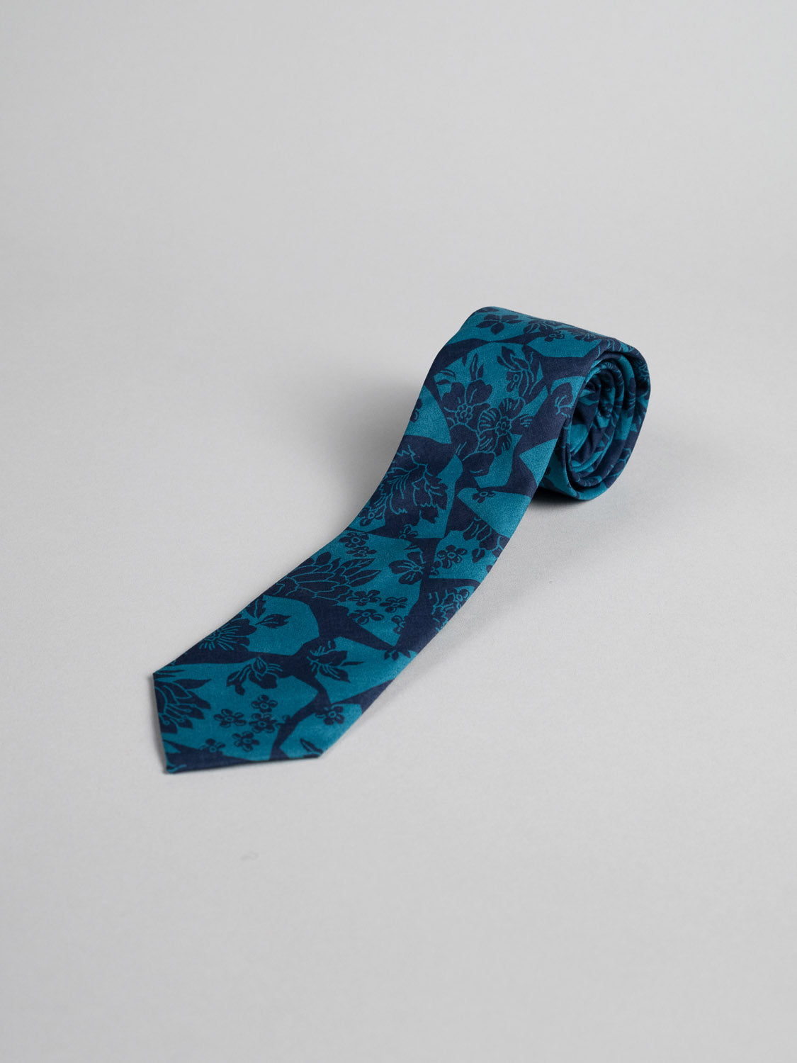 A Liberty of London Tana Lawn™ Ancient Tie by ID Menswear, handmade from teal cotton with a dark blue floral print, is neatly rolled on a plain light gray background.