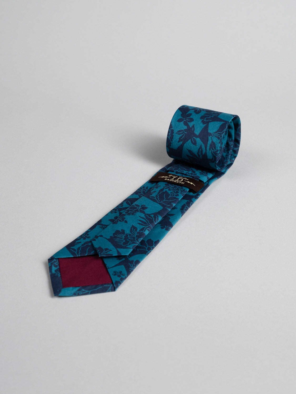The Liberty of London Tana Lawn™ Ancient Tie by ID Menswear—a teal tie with a dark blue floral pattern and maroon detail under the tip—handmade in Brooklyn, is rolled at one end and laid flat on a light gray surface.