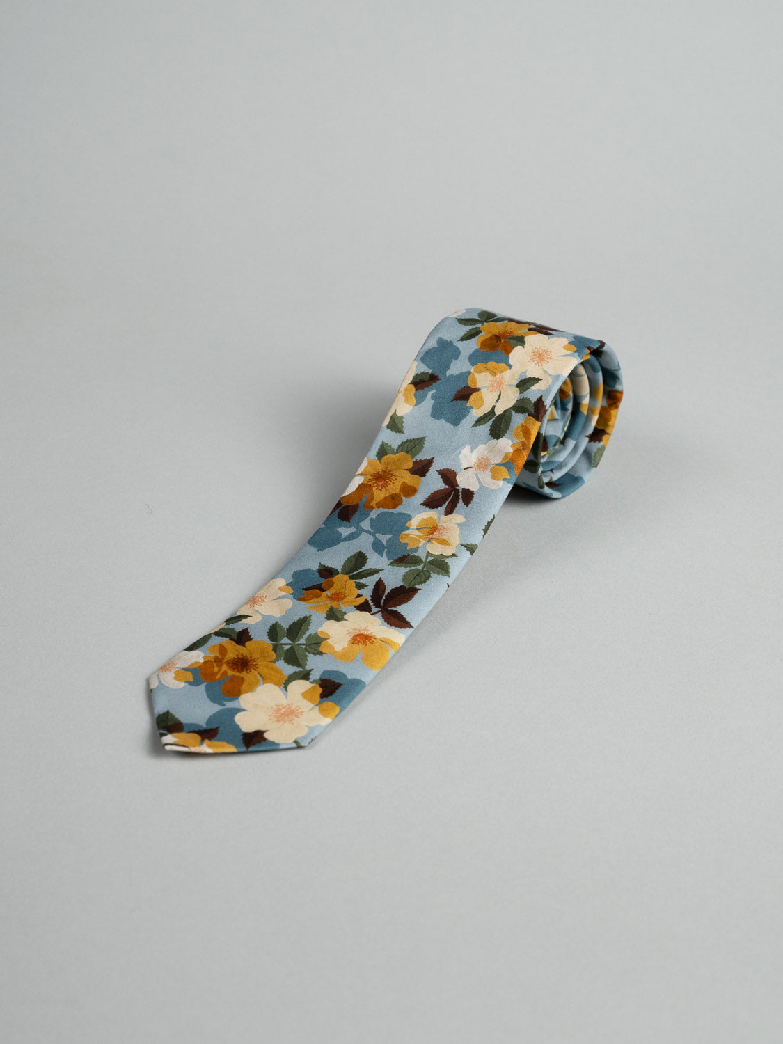 The ID Menswear Liberty of London Tana Lawn™ Nysa Tie, handmade in Brooklyn from luxurious cotton with a blue background and yellow, white, and brown floral pattern, is shown neatly rolled on a light gray surface.