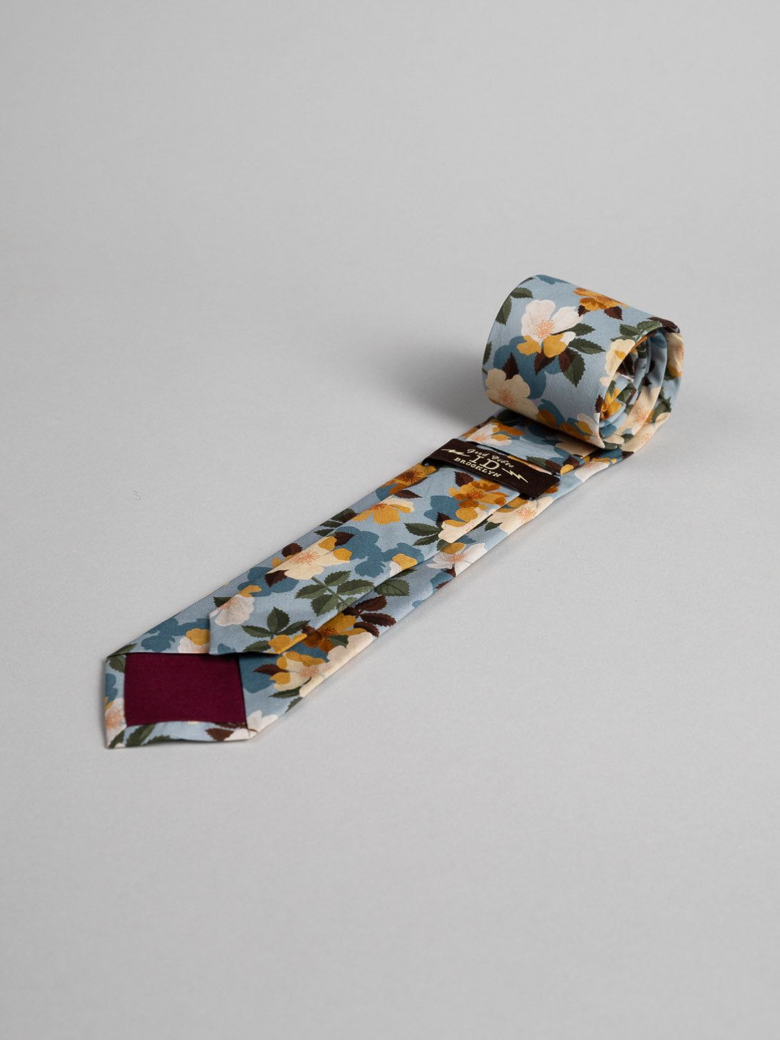 The Liberty of London Tana Lawn™ Nysa Tie by ID Menswear is handmade in Brooklyn from premium Tana Lawn cotton, featuring a blue, white, yellow, and brown floral pattern with a maroon tip, shown on a light grey background.