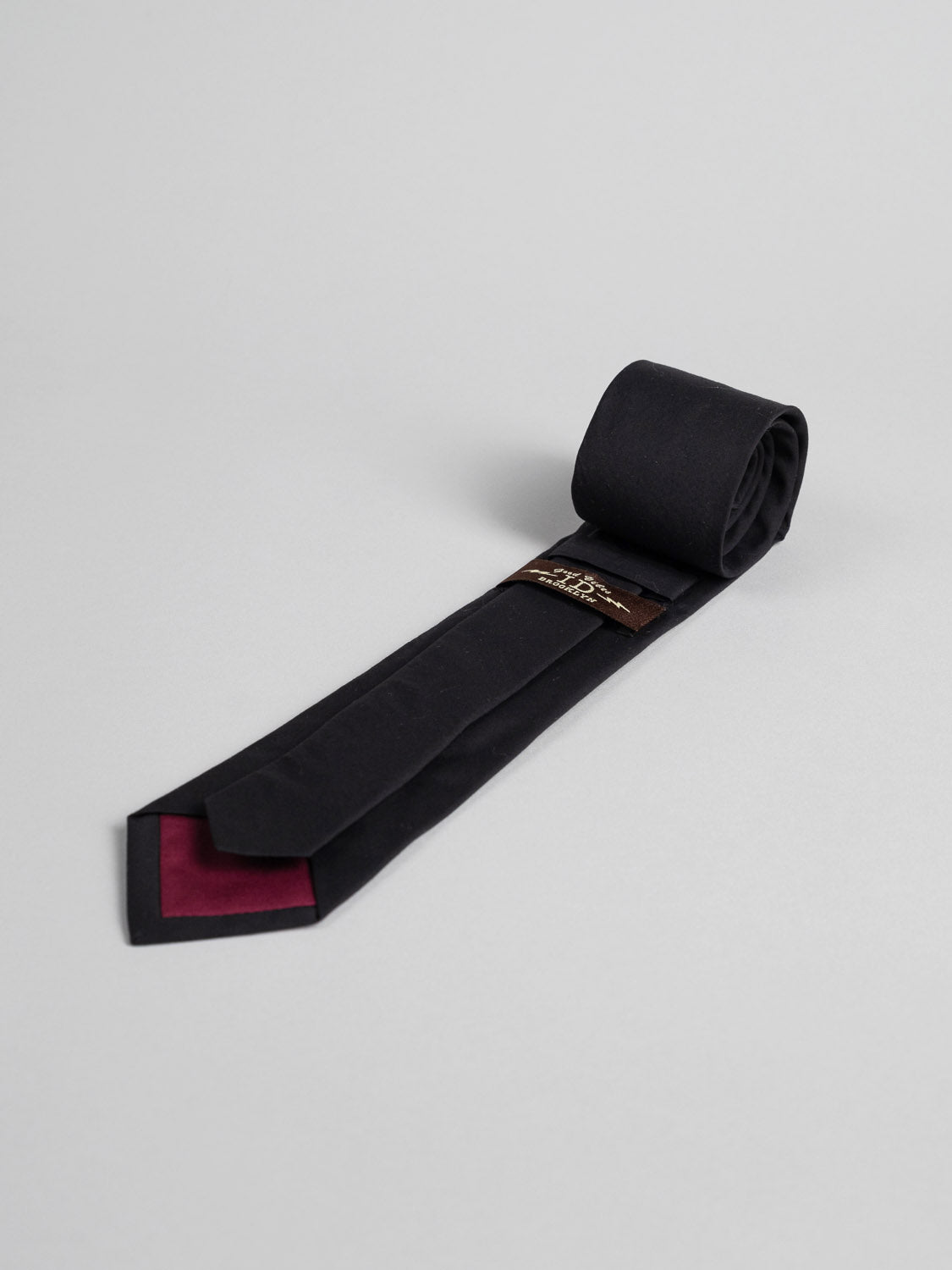 The Liberty of London Tana Lawn™ Noir Tie by ID Menswear is a black necktie, handmade and rolled on a light gray surface, featuring a maroon accent beneath the narrow end and a small brown label near the back.