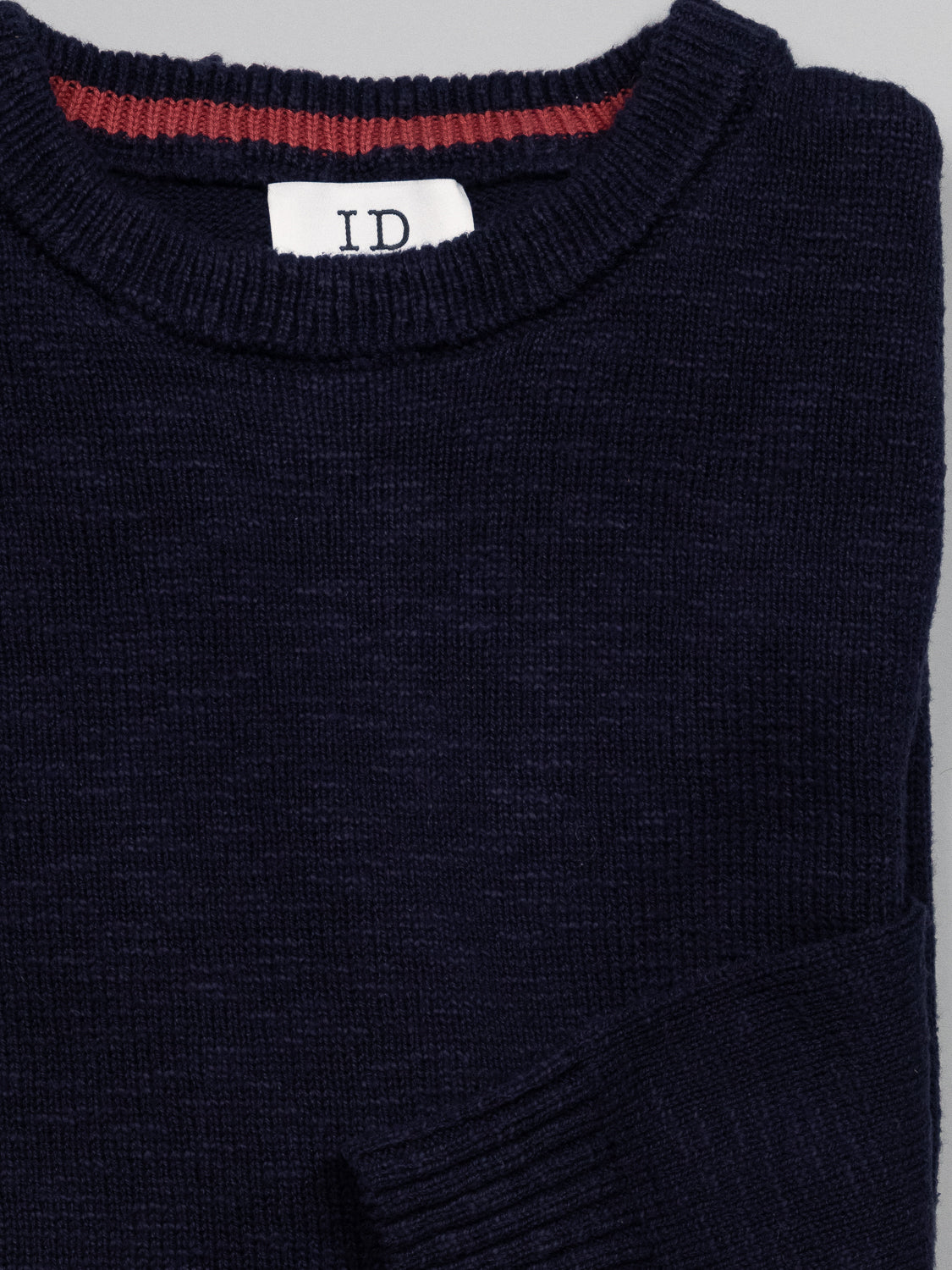 The Odense Slub Yarn Sweater by ID Menswear is a dark navy crew neck with a visible ID label inside. Made from slub cotton, it has a ribbed neckline with a subtle red stripe. This regular fit sweater is neatly folded against a plain, neutral background.