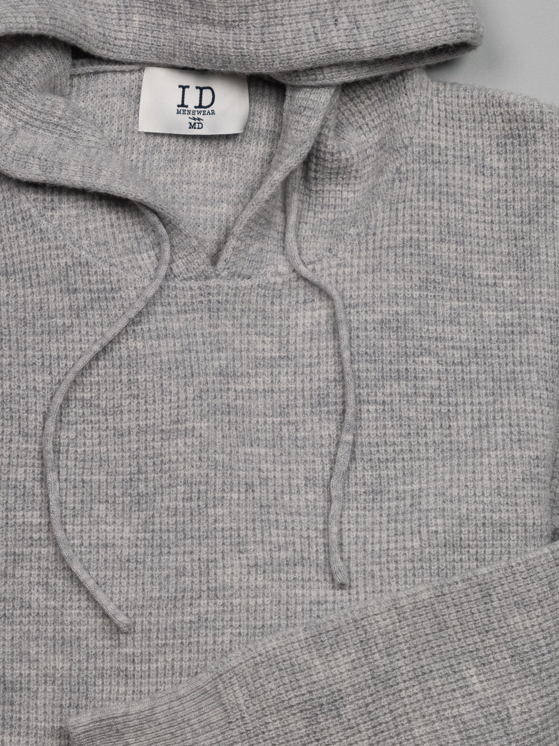 Close-up of a gray Bergen Waffle Knit Merino Hoody by ID Menswear, featuring a hood and drawstrings, with a white label reading ID Menswear inside the collar.