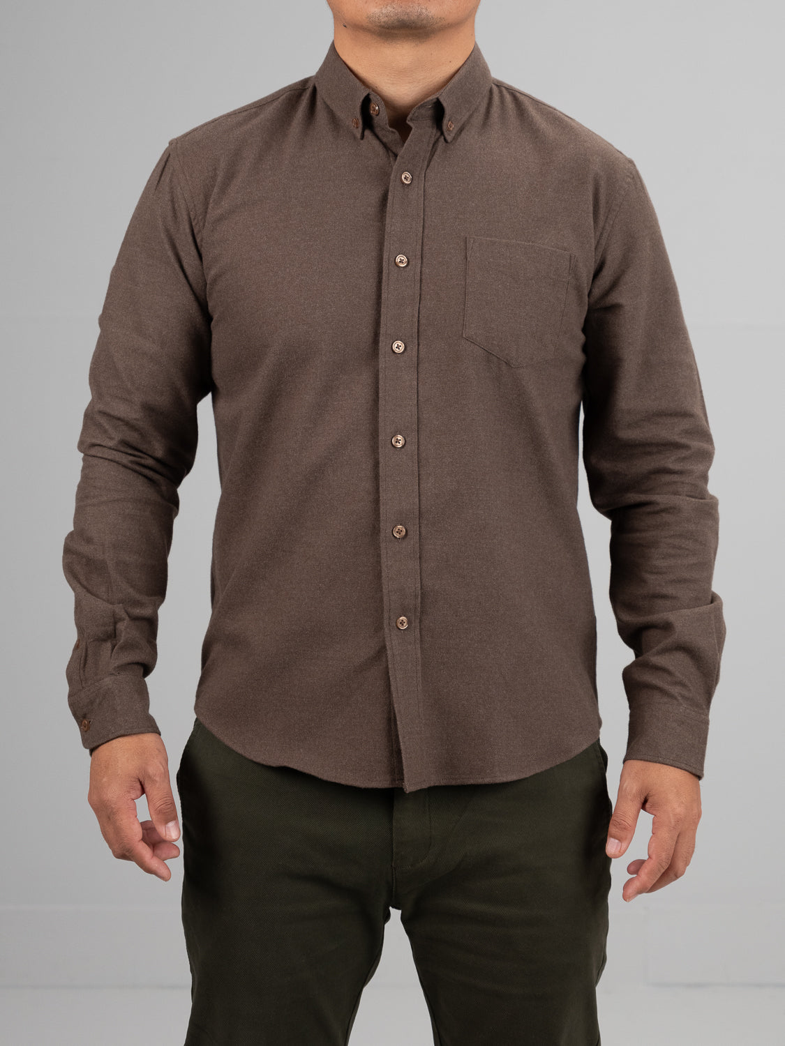 A person wears the Rex Cotton Sustans Blends Flannel Shirt by ID Menswear in brown—sustainably made with a chest pocket—paired with dark green pants, standing against a plain light gray background.