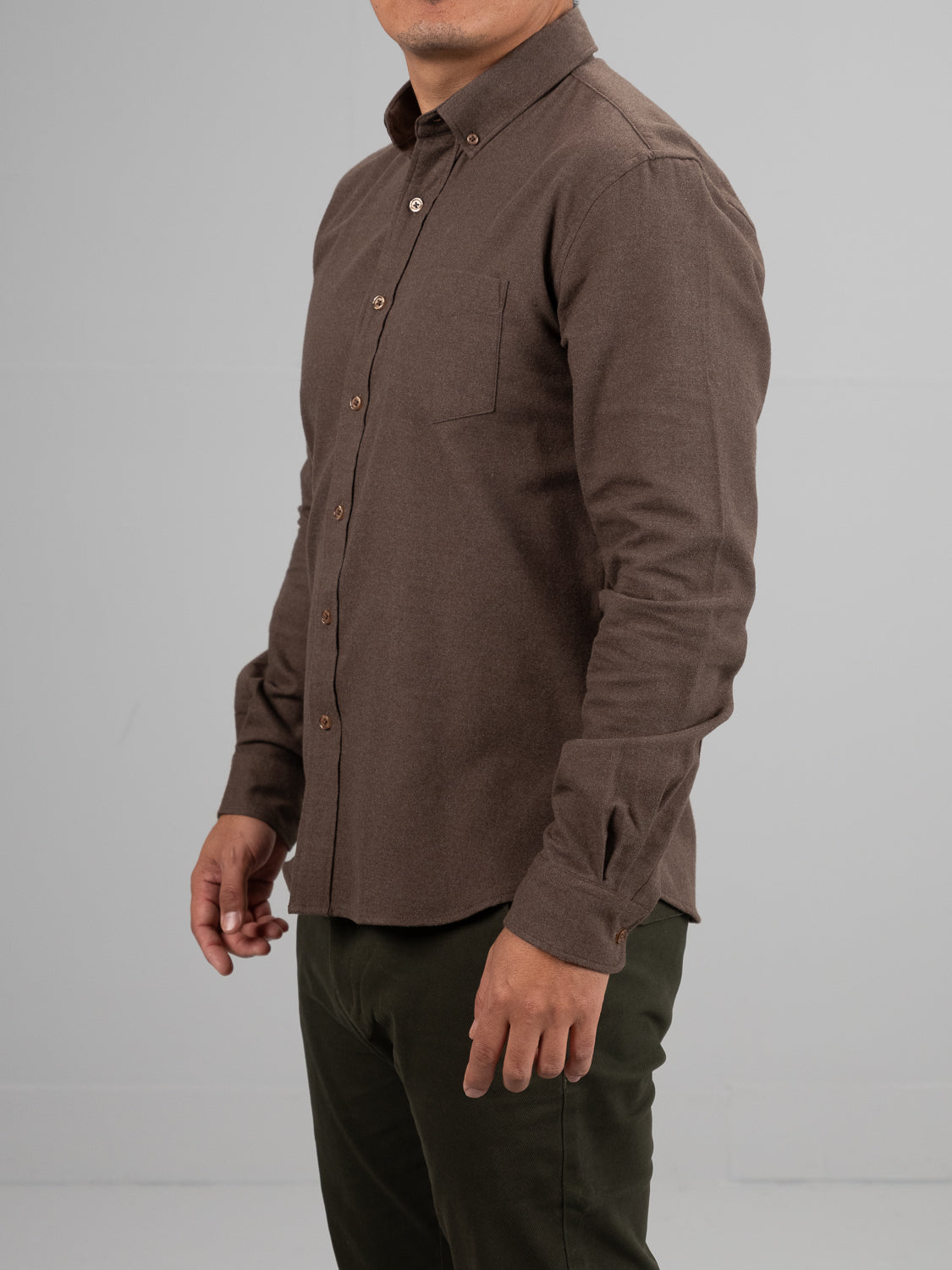 A man wears the Rex Cotton Sustans Blends Flannel Shirt by ID Menswear, an untucked brown long-sleeve button-up, paired with sustainable dark green pants, standing against a plain light gray background and facing slightly to the side.