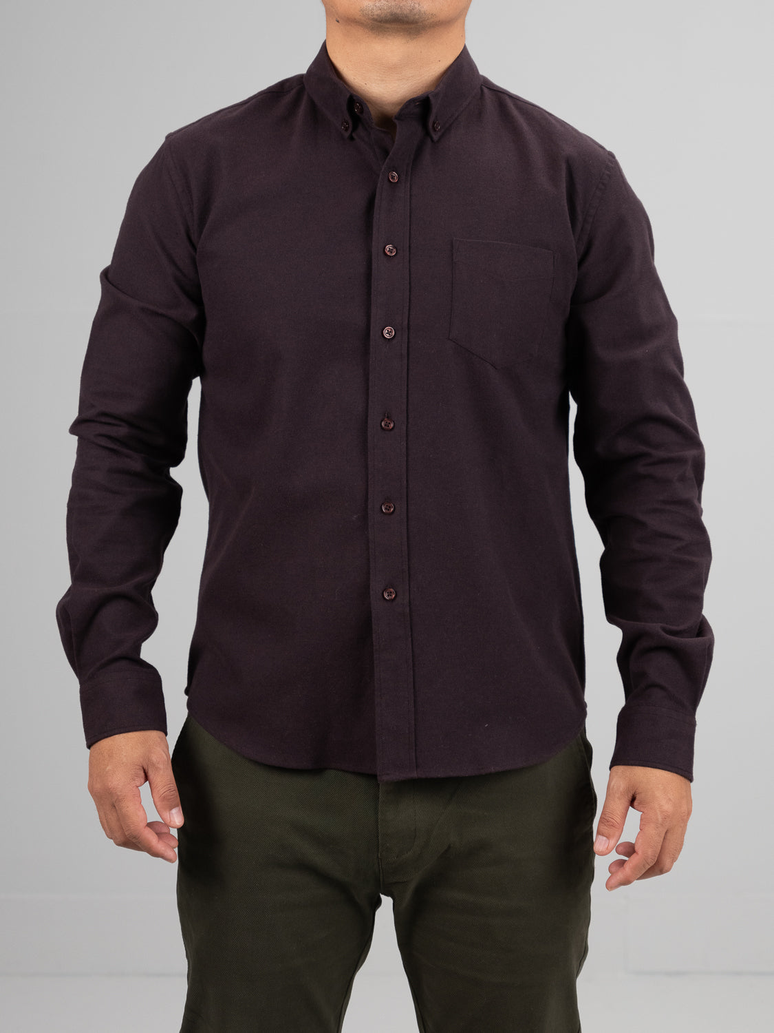 A person wears an ID Menswear Magnus Brushed Twill Solid Flannel Shirt, featuring a dark color, front pocket, and long sleeves, paired with olive green pants against a plain light gray background. The head is not visible in the image.