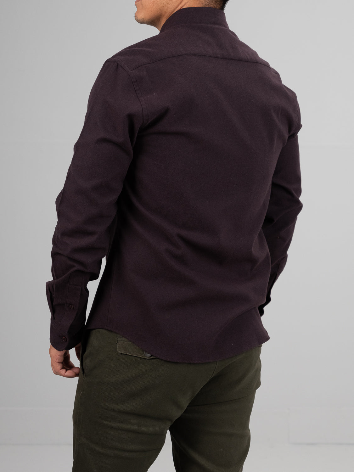 A person wearing an ID Menswear Magnus Brushed Twill Solid Flannel Shirt in dark brown and olive green pants stands with their back to the camera. The shirt is untucked, and their left arm is slightly bent against a plain light gray background.