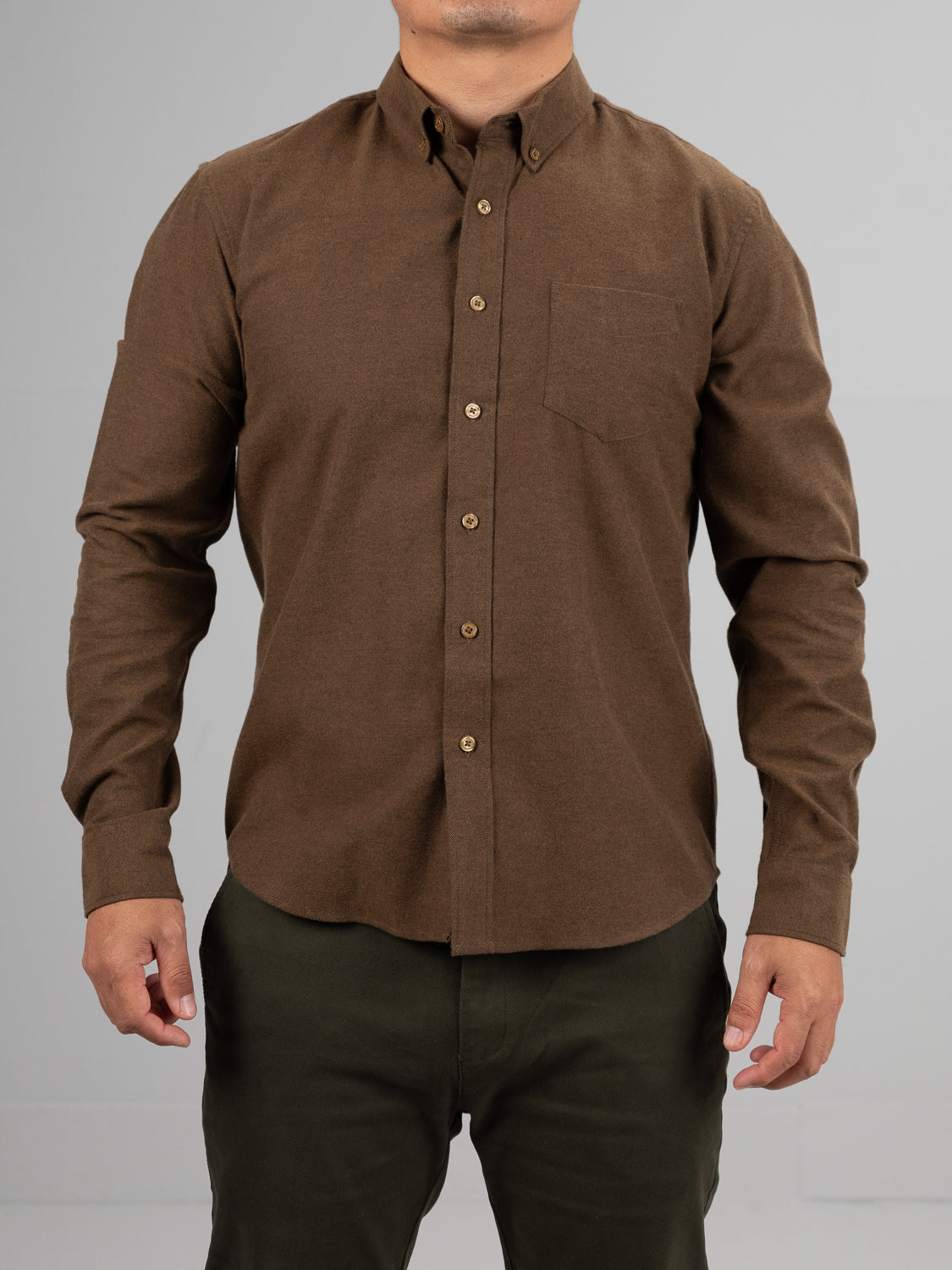 The person wears the ID Menswear Magnus Brushed Twill Solid Flannel Shirt in brown, featuring a button-down collar, chest pocket, and long sleeves, paired with olive-green pants. They stand against a plain gray background with their hands relaxed at their sides.